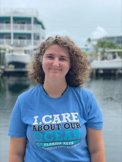 A woman with curly hair wearing a blue T-shirt that says 'I CARE ABOUT OUR OCEAN' standing in front of boats and a marina