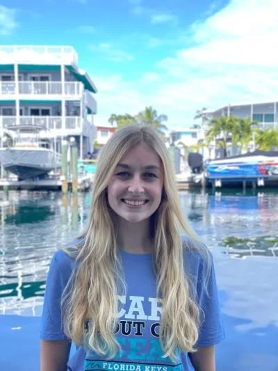 A young woman with long blonde hair smiling near a marina with boats and waterfront buildings in the background.