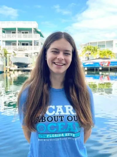 Young woman with long brown hair smiling at the camera, standing by boats and waterfront buildings under a blue sky.