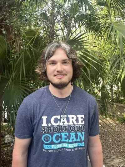 A young man with wavy, grayish hair and a beard stands outdoors among lush green palm plants. He is wearing a navy blue T-shirt that reads 'I CARE ABOUT OUR OCEAN FLORIDA KEYS' and a silver necklace.