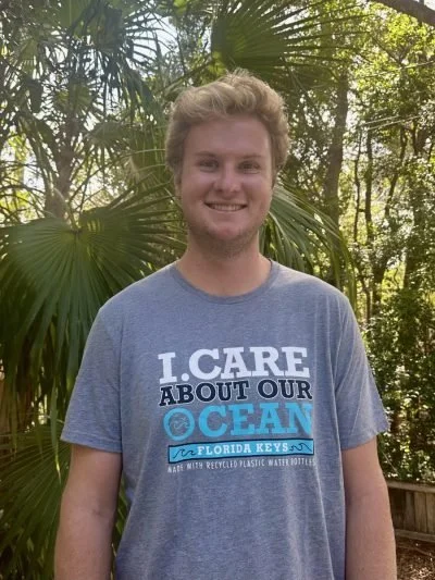 Young man standing outdoors in front of tropical trees, smiling, wearing a gray T-shirt promoting ocean conservation.