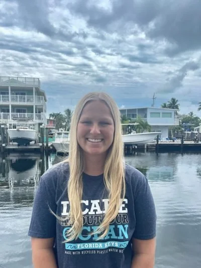 Young woman with long blonde hair smiling outdoors near a marina with boats and buildings under a cloudy sky.