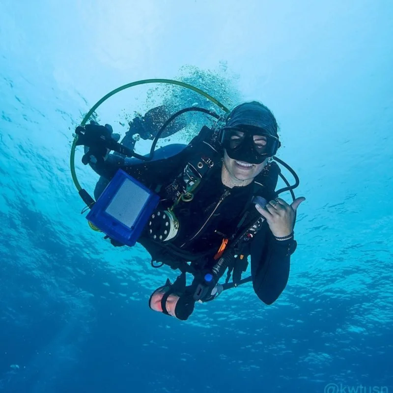A woman scuba diving underwater, smiling, wearing a wetsuit, mask, and holding a signaling device, with a blue and turquoise ocean background.