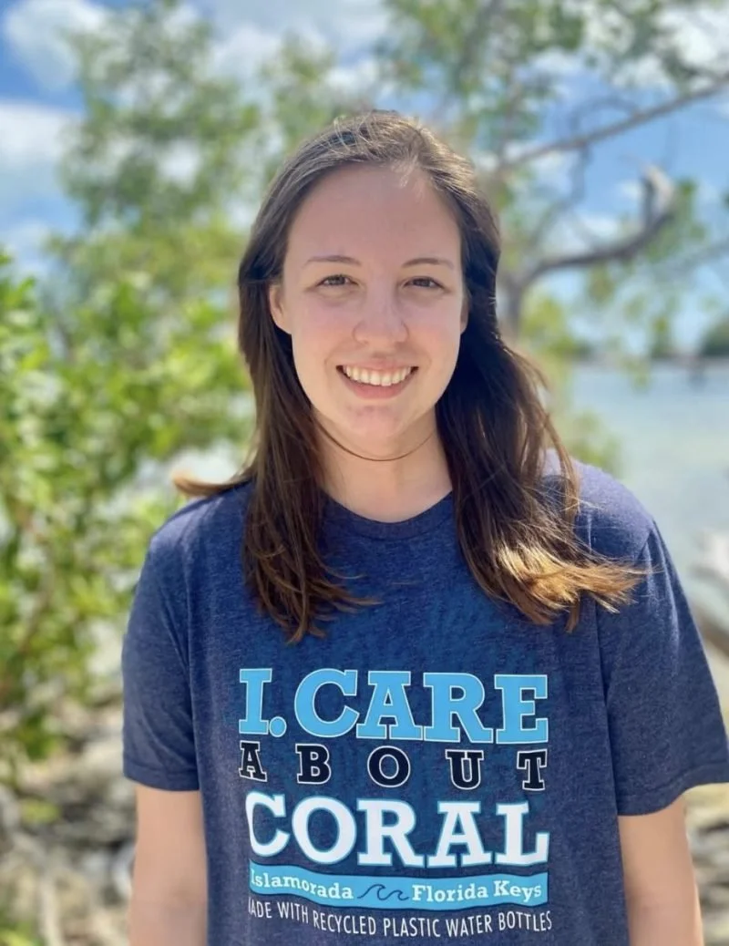 Smiling young woman with brown hair wearing a blue T-shirt that reads "I care about coral" and has logos for "Islamorada Florida Keys" and mentions recycled plastic water bottles, standing outdoors near water and greenery.