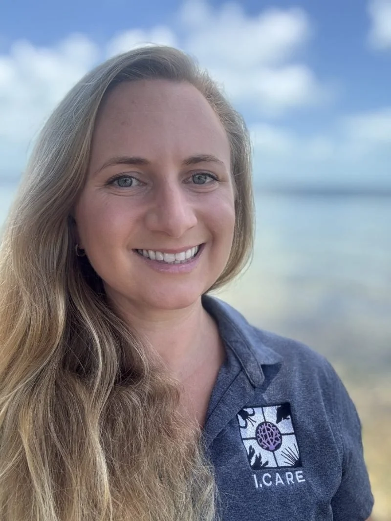 A woman with long blonde hair smiling at the camera, wearing a dark gray polo shirt with a logo that says I.CARE, standing outdoors near the water with a cloudy sky in the background.