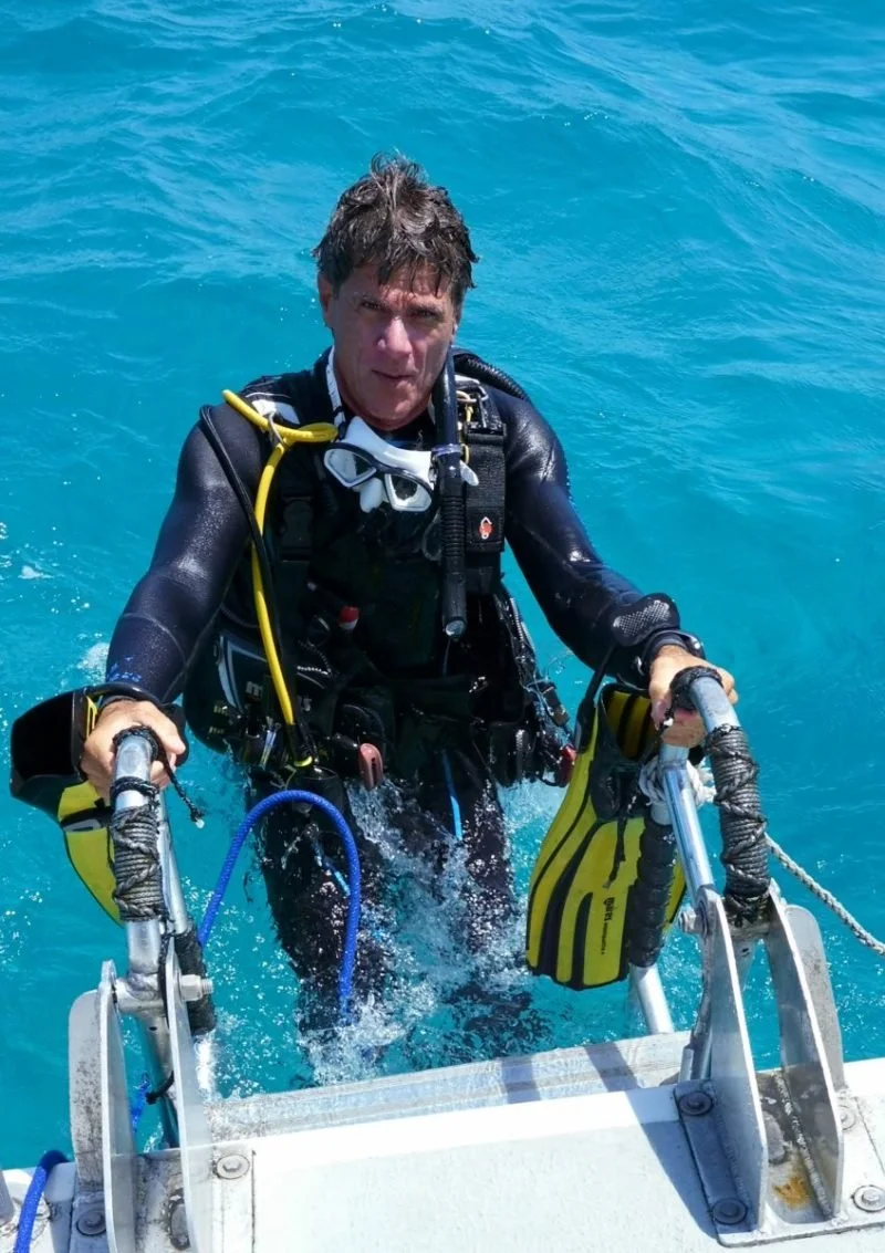 Person in scuba diving gear climbing out of the water on a metal ladder attached to a boat, with blue ocean in background.