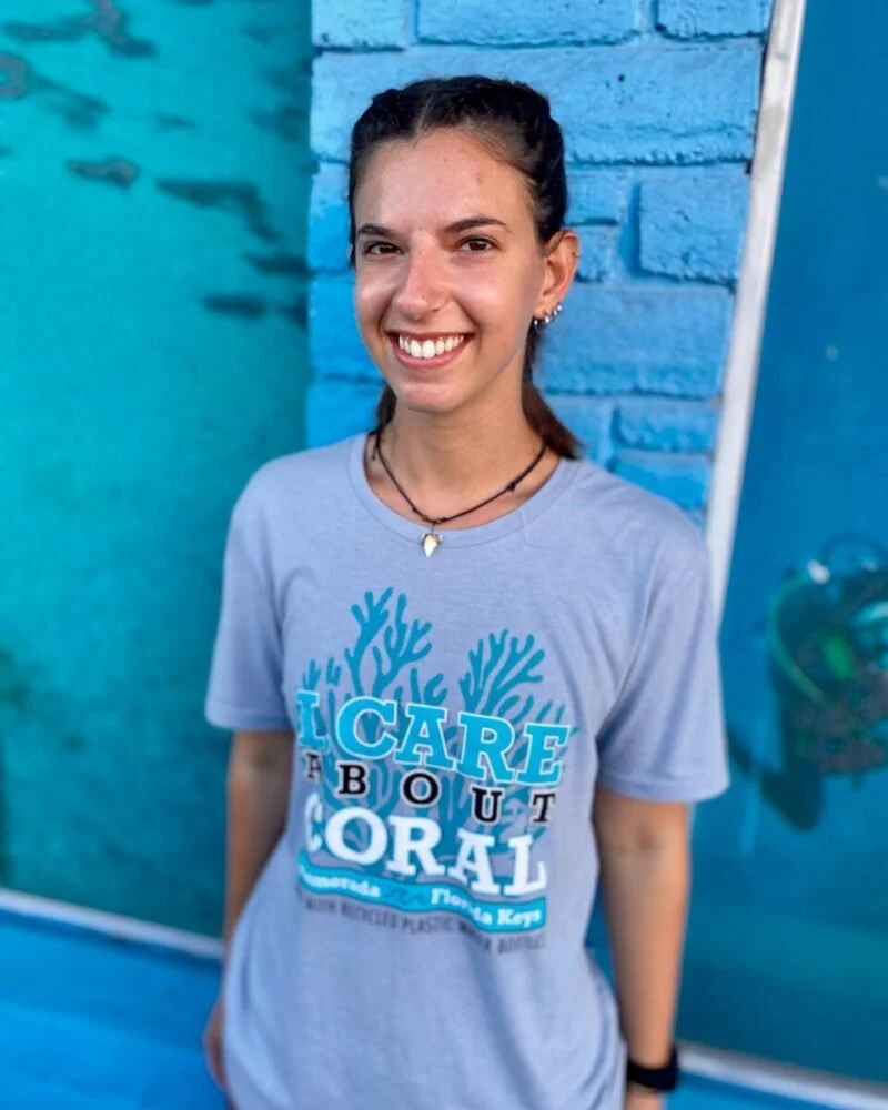 A young woman with dark hair tied back, wearing a gray T-shirt with an ocean-themed design and text, standing in front of a blue wall with a coral reef image and smiling at the camera.