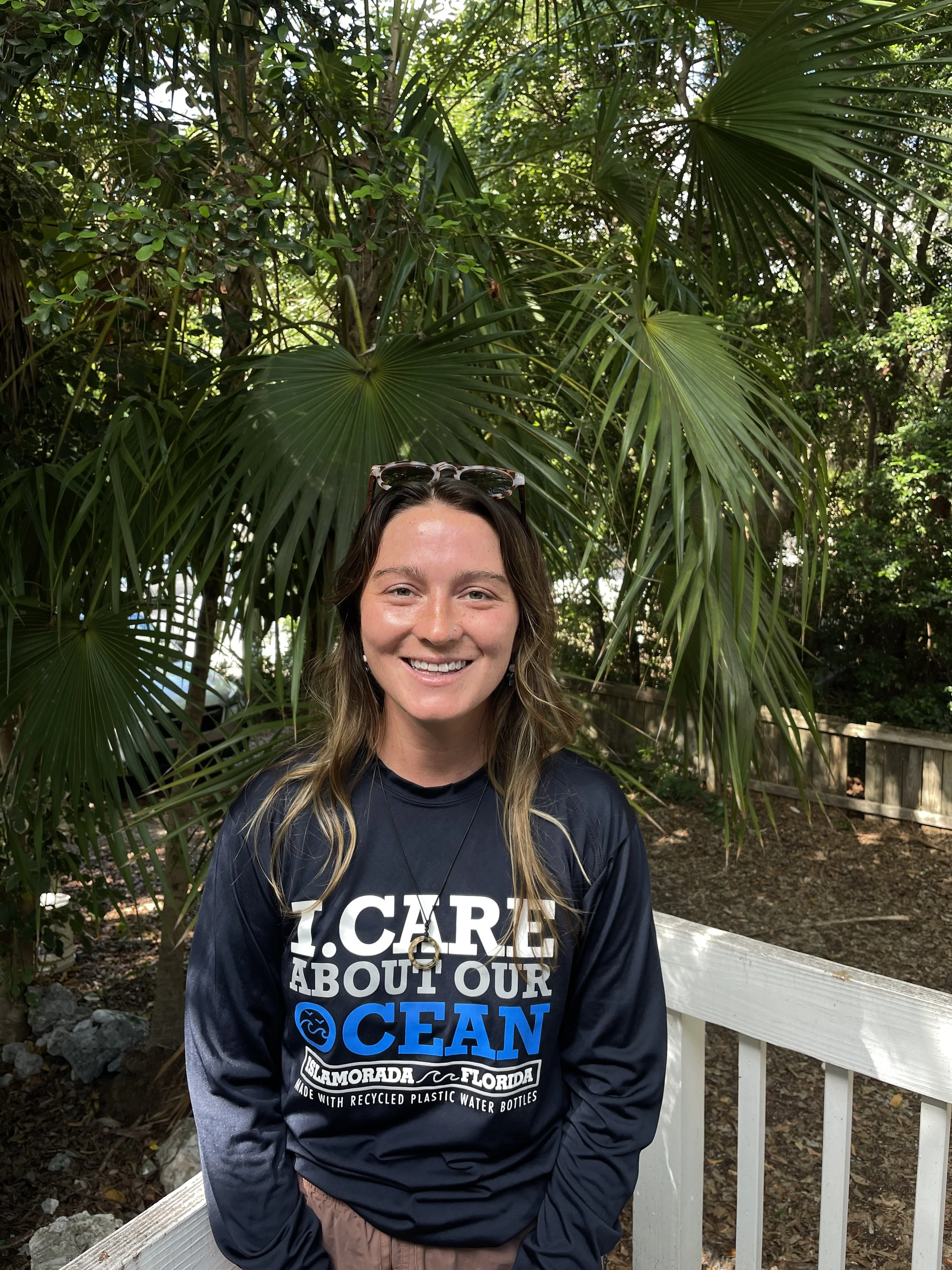 A woman with long brown hair, smiling, wearing sunglasses on her head and a black long-sleeve shirt that reads 'I.CARE ABOUT OUR OCEAN' with additional text referencing Islamorada, Florida, standing outdoors in front of tropical plants and a white wooden fence.