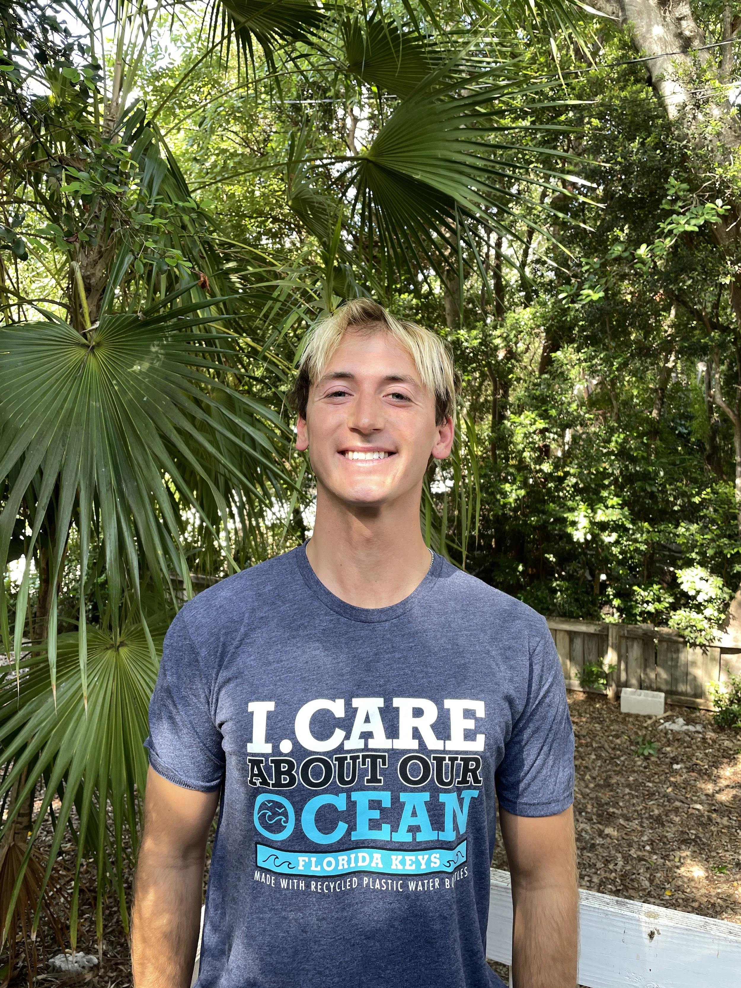 A young man with blonde hair and a gray t-shirt smiling outdoors, surrounded by green trees and plants.