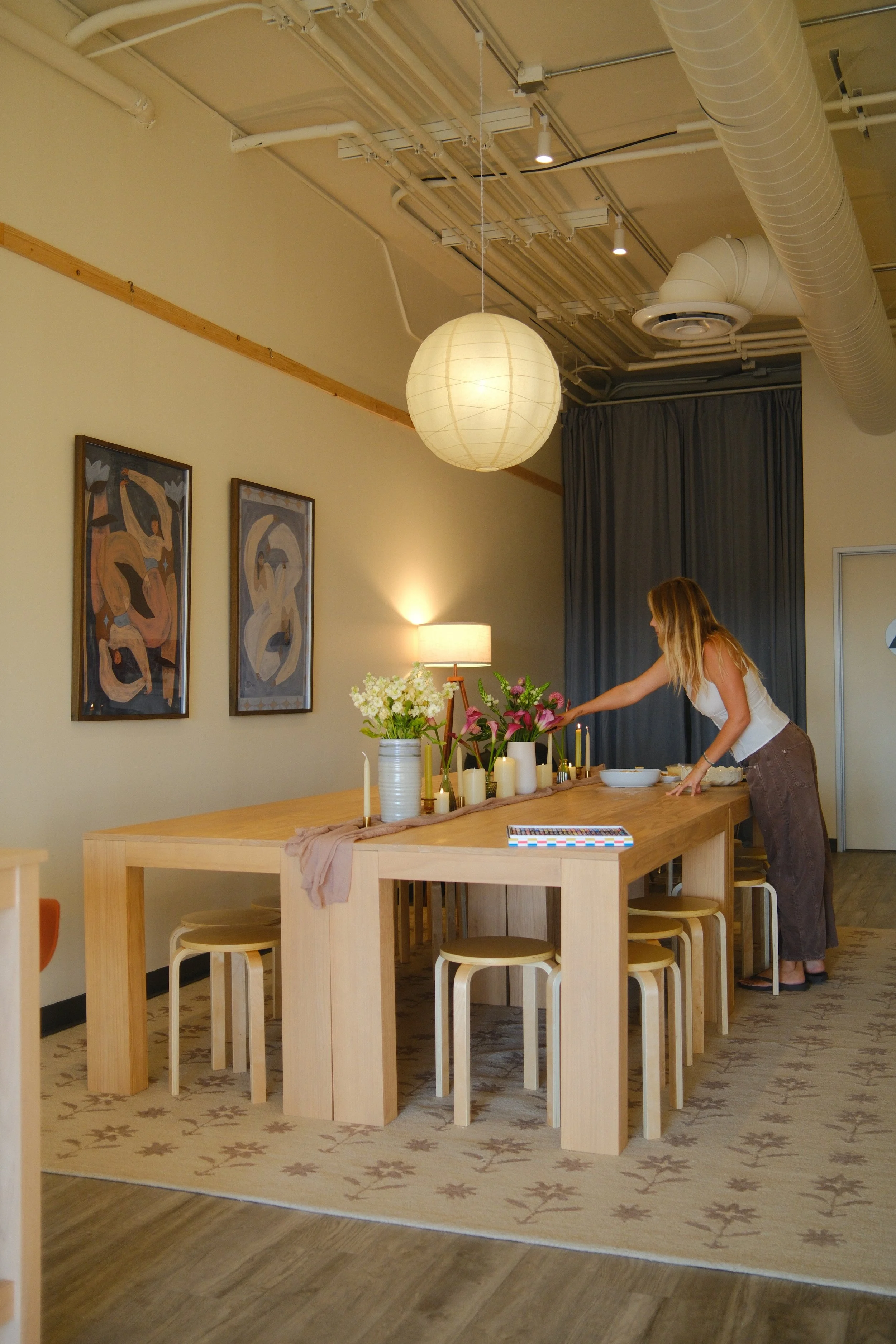 A woman is arranging flowers on a wooden dining table decorated with candles and vases. The table is in a room with hanging artwork, a paper lantern ceiling light, and industrial-style ceiling ducts.