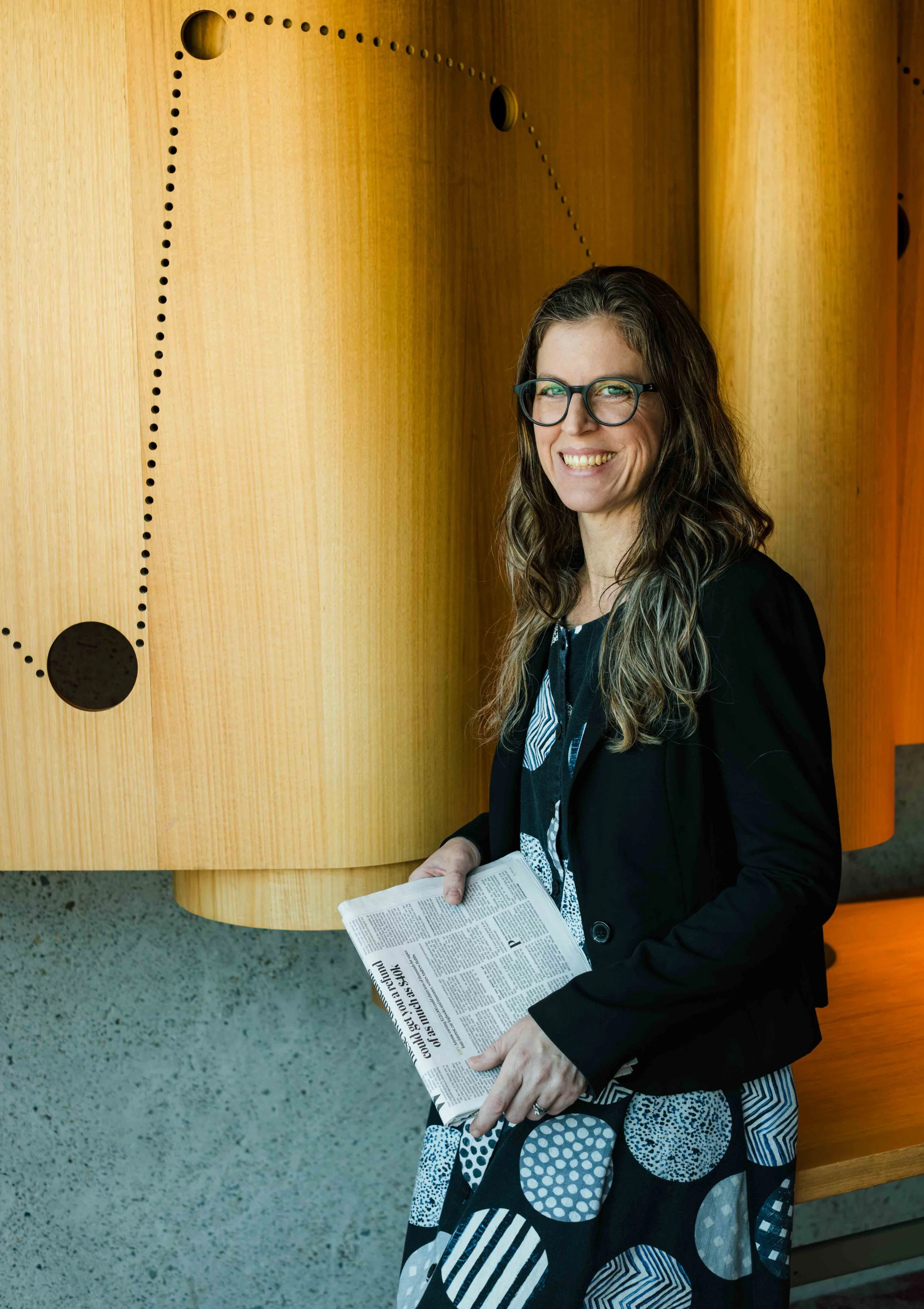 A female lawyer with long, wavy brown hair and glasses, smiling, holding a newspaper, standing next to a wooden, curved exhibit display with circular holes.