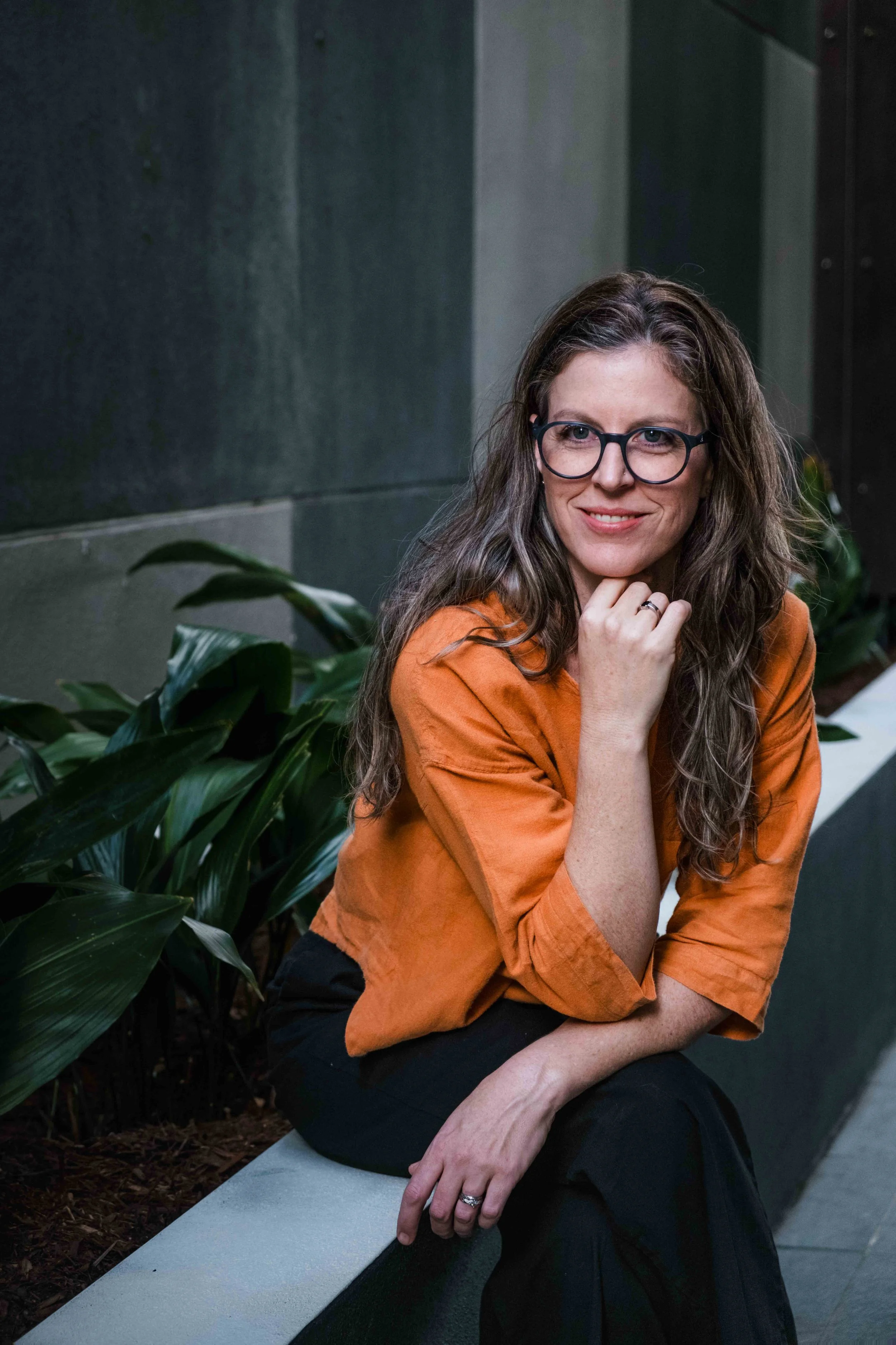 A female lawyer with curly brown hair wearing black glasses and an orange top sitting on a concrete bench outdoors, surrounded by green plants, smiling and resting her chin on her hand.