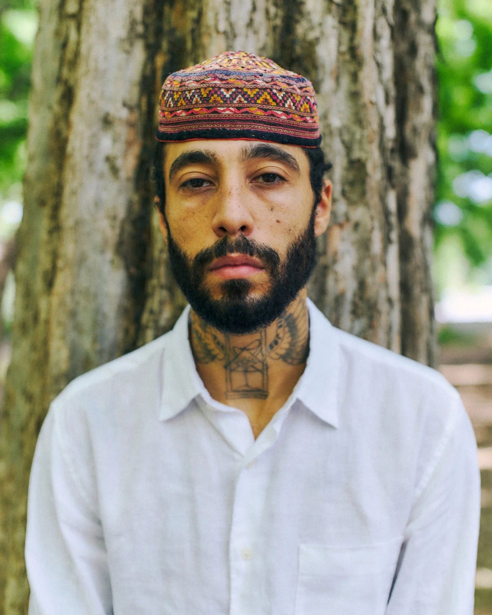 A man with a beard, tattoos on his neck, and a traditional embroidered cap, standing in front of a tree with green foliage.