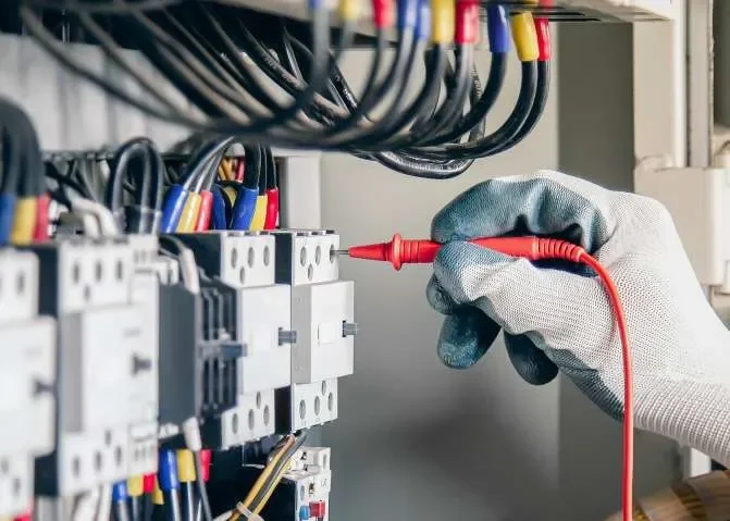 A technician's hand wearing a gray glove holding a red test probe while working on electrical circuit breakers with multiple colored wires.