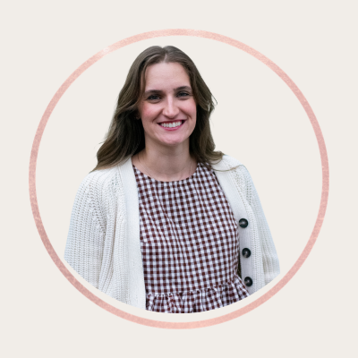 A woman smiling, wearing a checkered dress and a white cardigan, against a light background with a pink circular border.