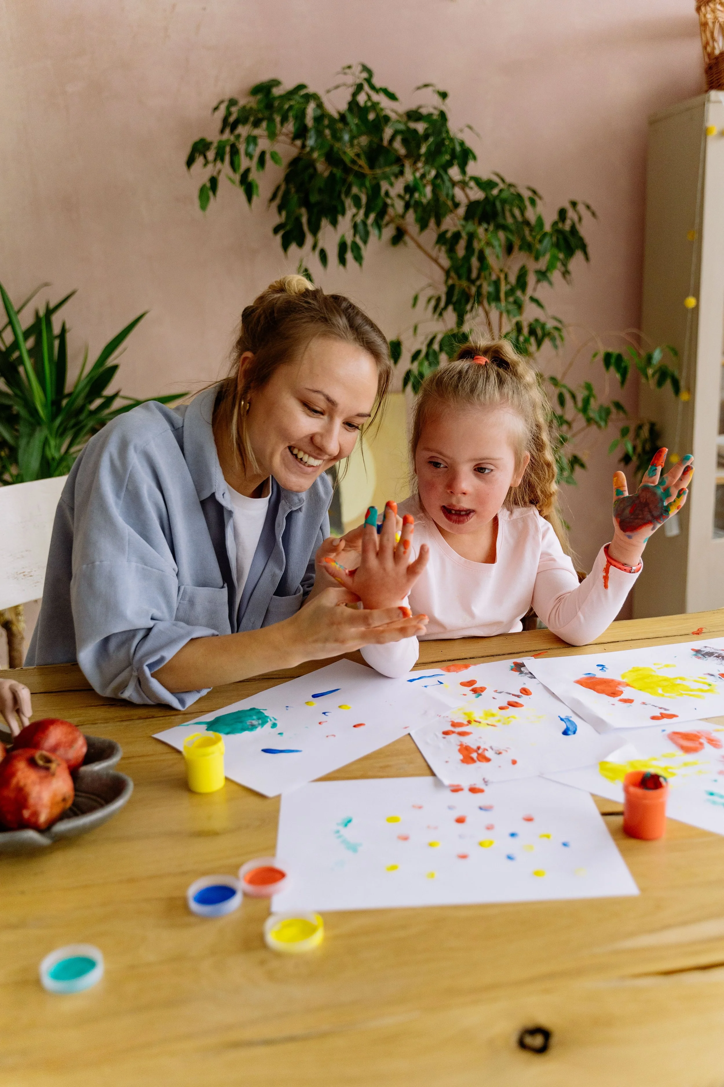 A woman and a young girl are sitting at a table, engaging in painting activities. The woman is smiling and showing the girl her painted hand, which she is holding up. The table is covered with sheets of paper with colorful paint splatters, and there are small containers of paint in various colors. In the background, there are green plants and a beige wall.