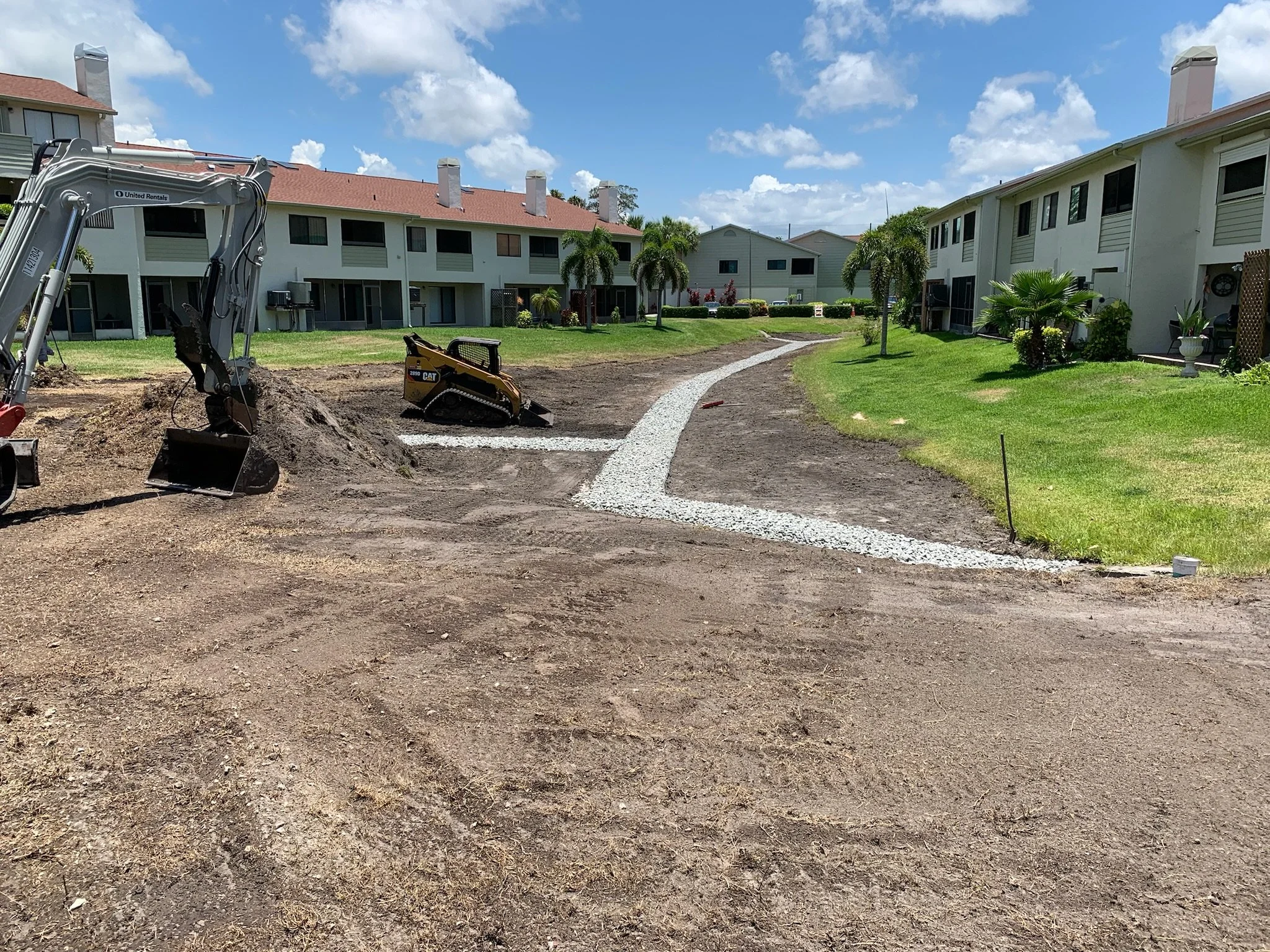 Apartment complex's courtyard with ongoing landscaping work, showing a partially finished gravel pathway, mechanical equipment, and lush green grass and palm trees.