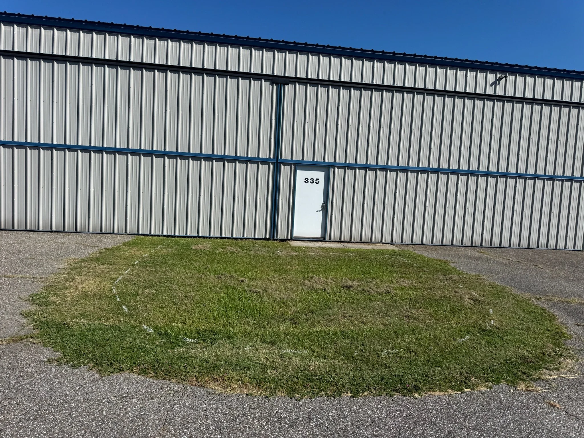 A large gray metal building with a white door labeled '335' and a small green grass patch in front. Vegetation removal prep.