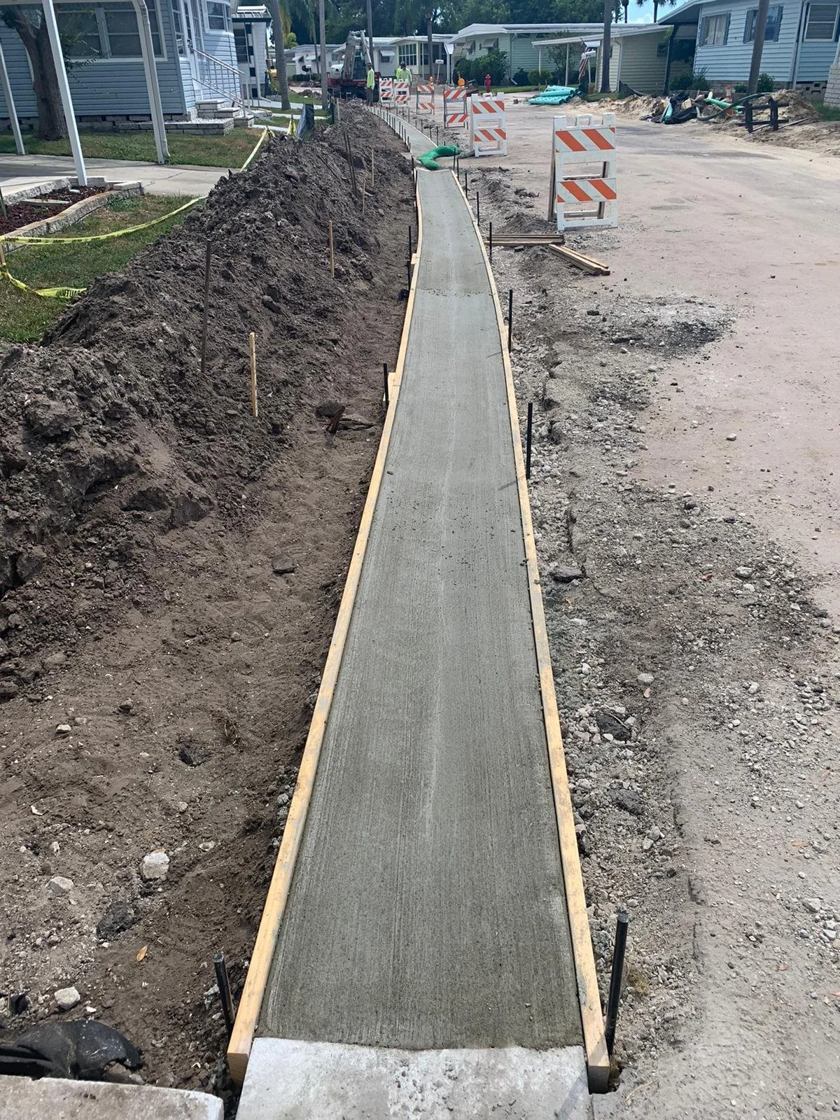 Newly poured concrete curb under construction in a residential neighborhood with orange and white barricades, construction equipment, and workers in the background.