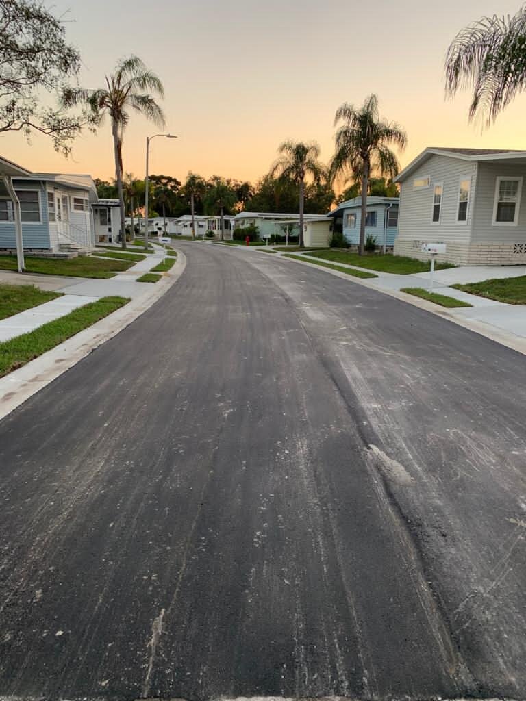 A quiet suburban street at sunset with dirt in the middle of the paved road, surrounded by small houses with lawns and palm trees. Finished roadway reconstruction.