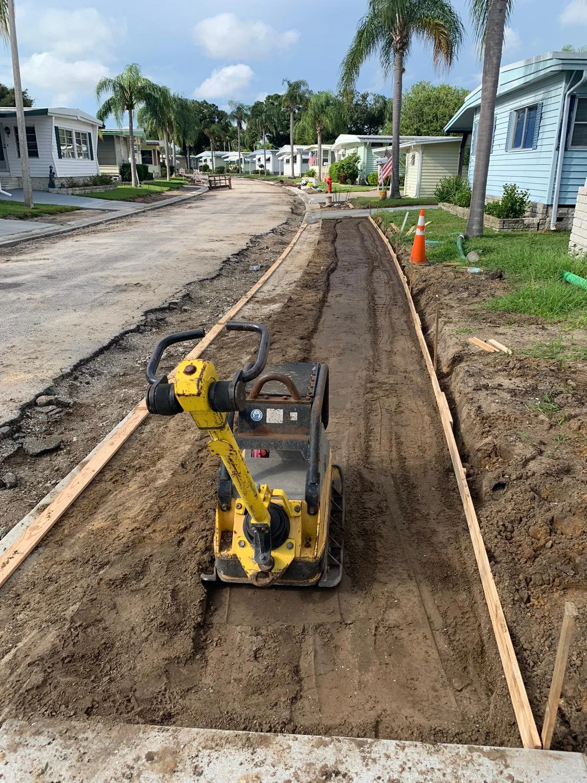 A construction site on a residential street shows a pathway being prepared with wooden frames for concrete pour, with a yellow compactor machine in the foreground, surrounded by houses and palm trees under a partly cloudy sky.