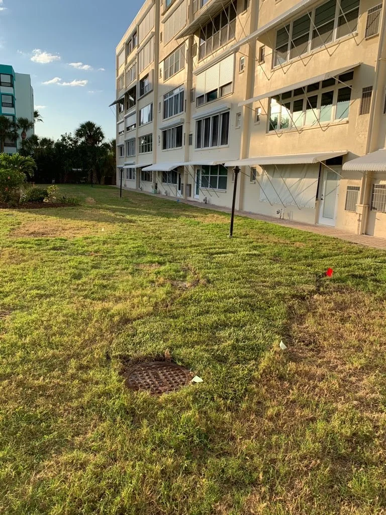 Green grassy lawn with a drain cover in the foreground, surrounded by patchy grass, next to a multi-story beige apartment building with windows, balconies, and awnings, under a blue sky with some clouds.