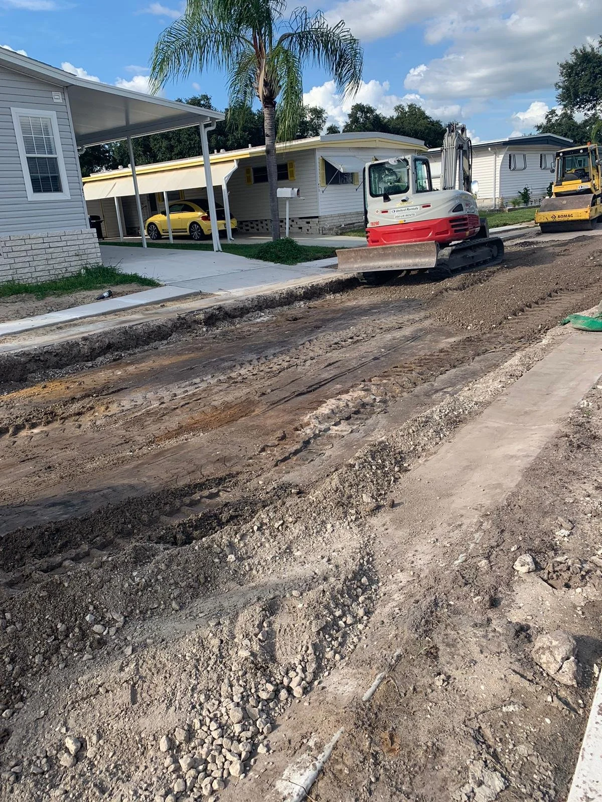 A construction site in a residential neighborhood with a dirt road undergoing roadwork, construction equipment, and houses with parked cars in the background. Roadway reconstruction.