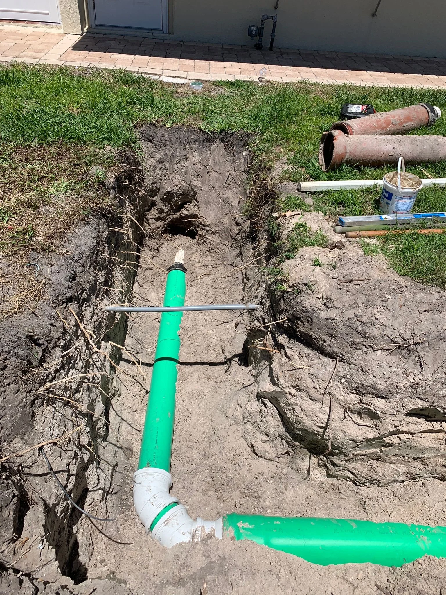 Underground piping installation with green and white pipes connected with fittings, pipes are in a trench with exposed soil and roots, near a house with a brick walkway and grass.