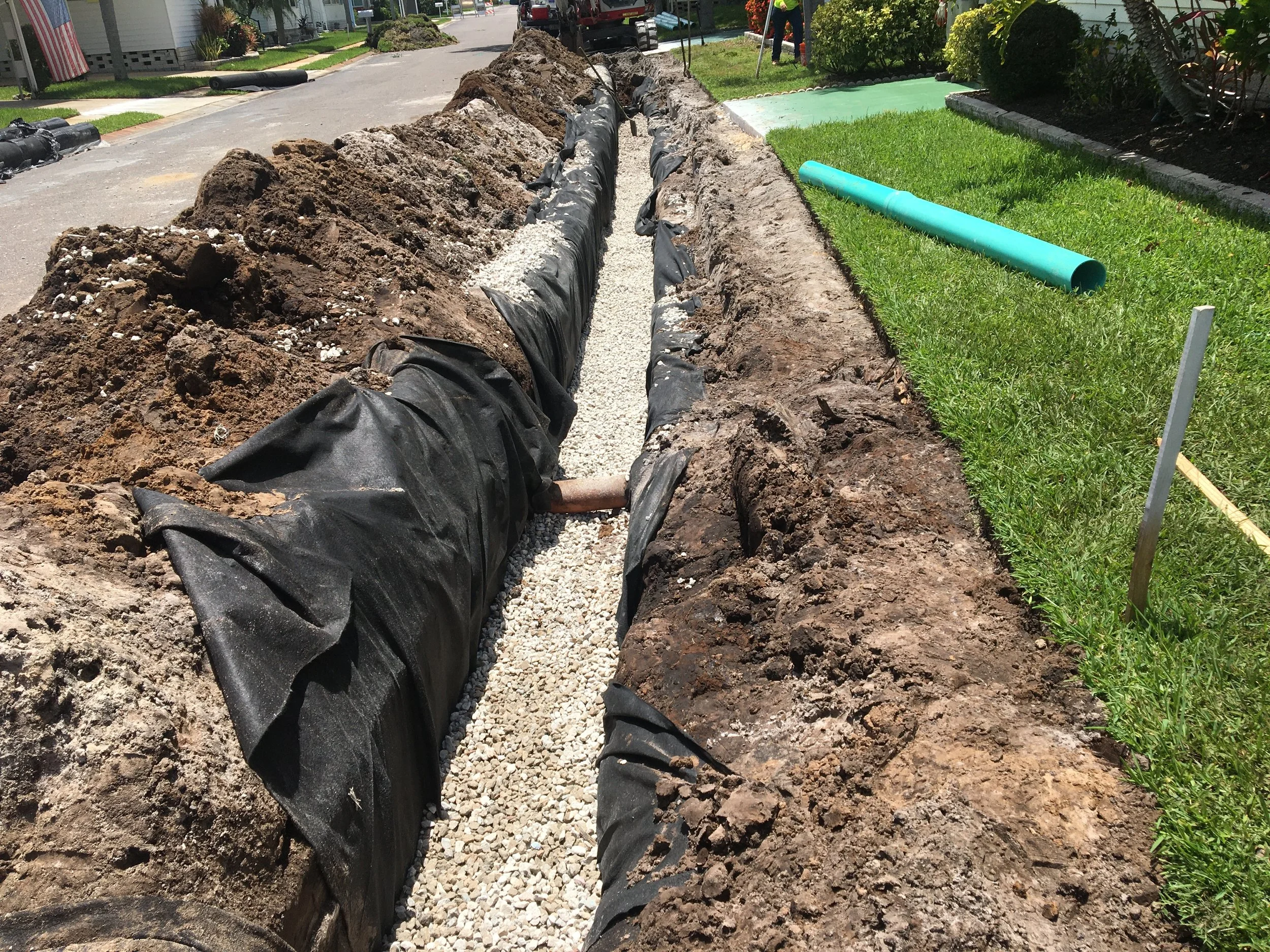 Construction site on a residential street with a trench being dug. Black landscaping fabric lines the trench, and gravel is being added inside. A green pipe is on the grass nearby. Houses and trees are in the background. Underdrain installation.