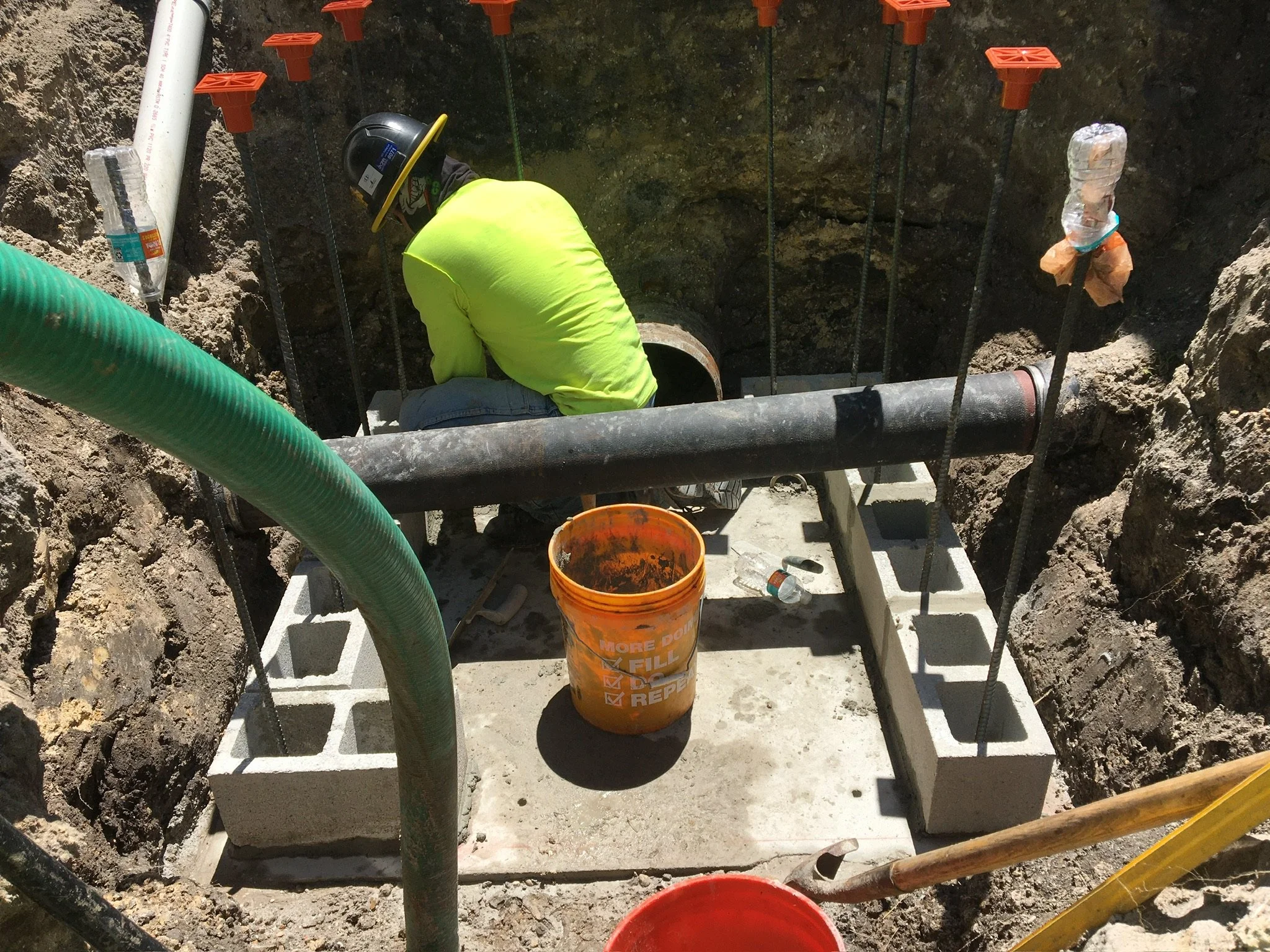Worker wearing a yellow safety vest and helmet working inside an excavated trench with utility pipes and a white concrete foundation on the ground.  Utility conflict structure.