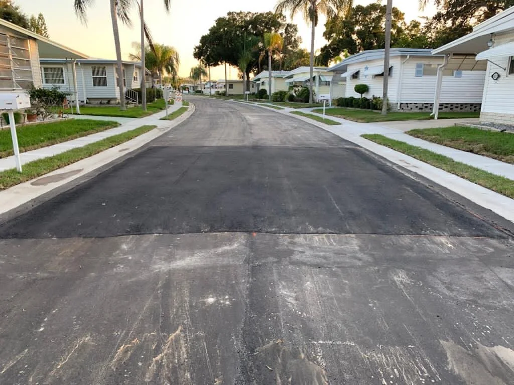 Residential street with newly paved blacktop, white concrete sidewalks, grassy lawns, palm trees, and single-story white houses with covered porches.