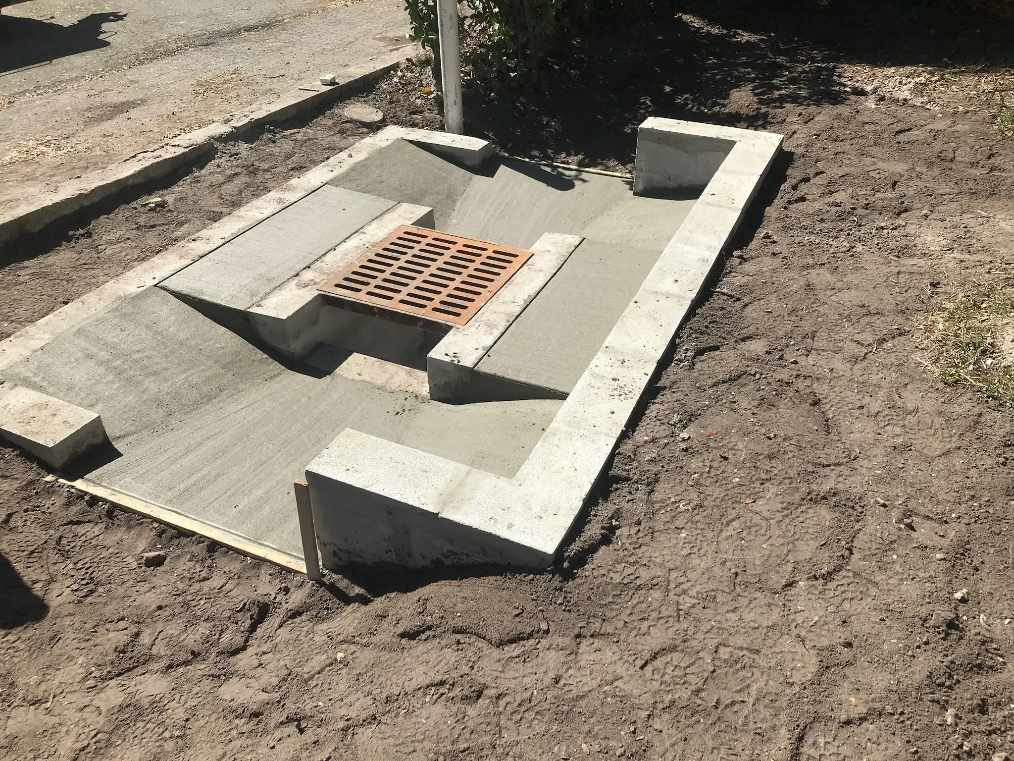 A newly constructed concrete sidewalk with a drainage grate, surrounded by dirt and Soil, and some construction materials nearby.