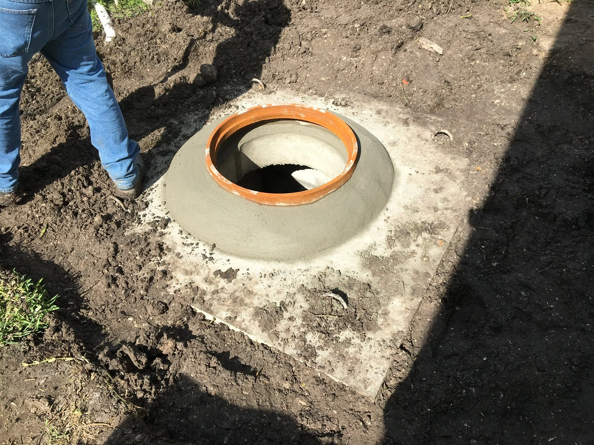 A construction worker in blue jeans standing next to a newly installed pipe in the ground, surrounded by dirt and soil. Utility conflict structure top an manhole lid.