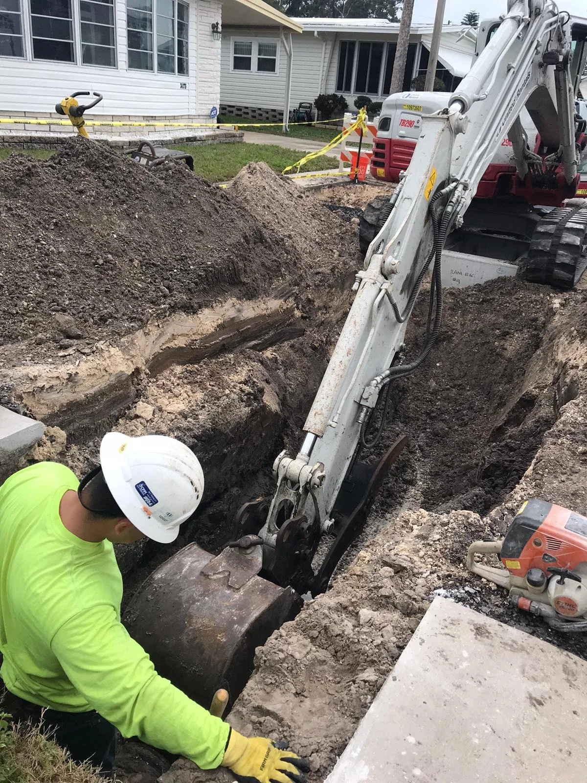 Worker in a neon yellow shirt and white helmet working in a large excavated trench with construction equipment around, including an excavator and ground tools, in a residential neighborhood.