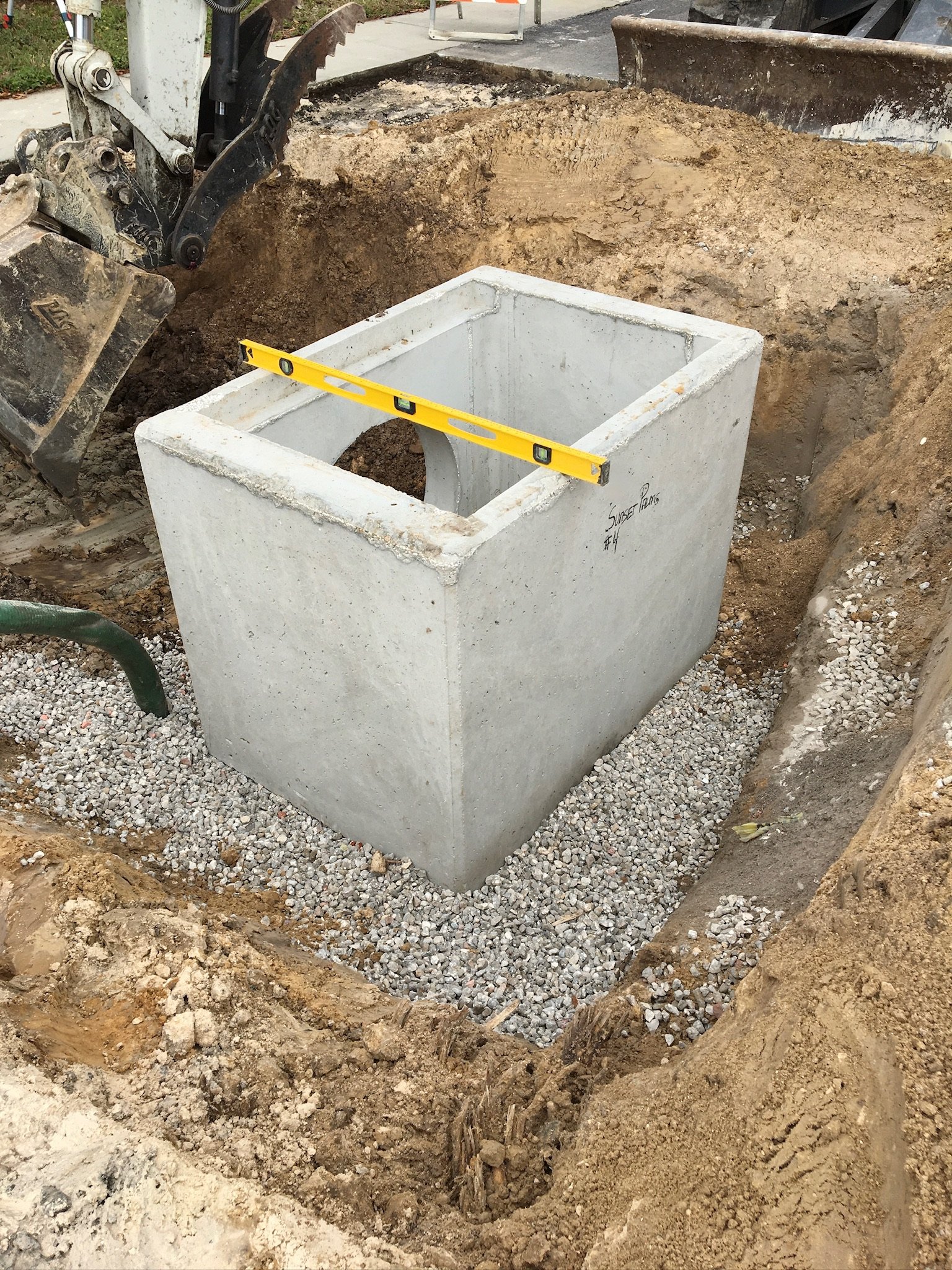 A construction site showing a concrete utility box inside an excavation with dirt and gravel surrounding it. A level tool is placed across the top of the box.