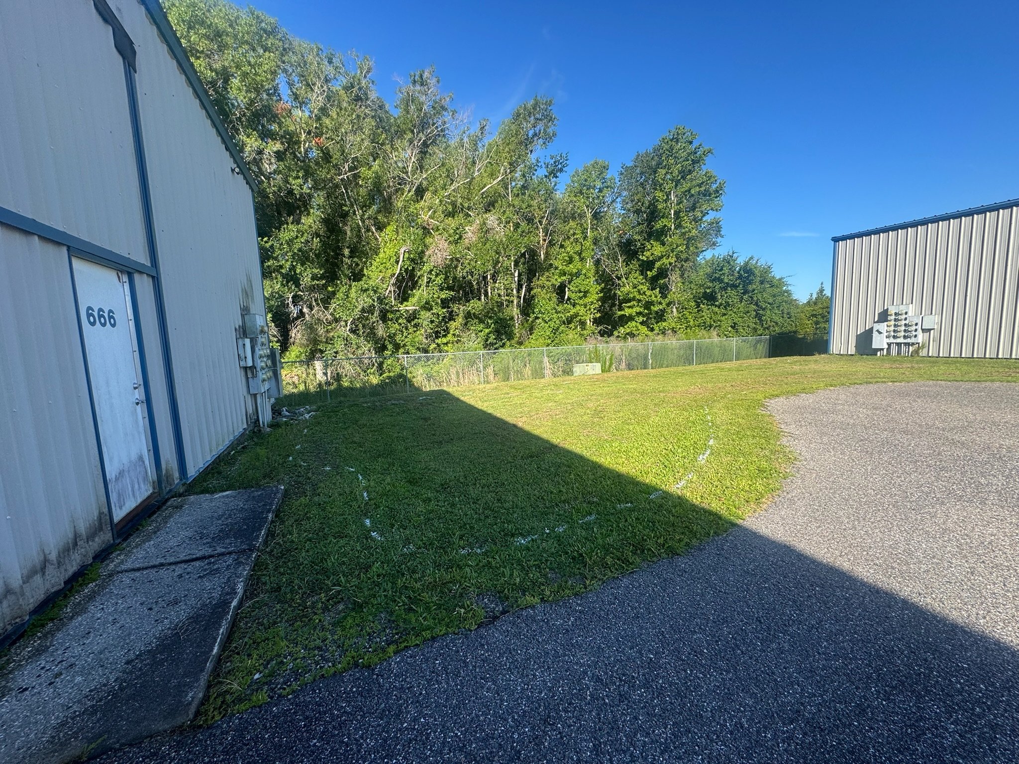 View of a building with metal siding and a grassy area, a chain-link fence, and a backdrop of green trees under a clear blue sky. Vegetation removal prep.