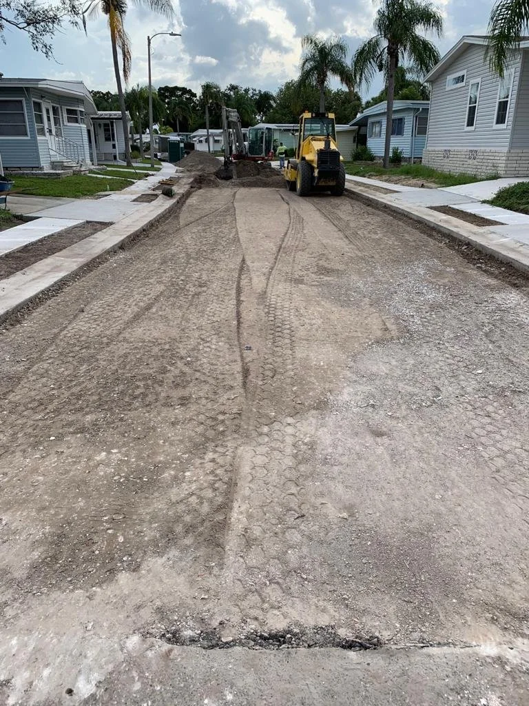 A residential street under construction with heavy machinery, including a yellow bulldozer and an excavator, working on road repairs. Sidewalks are in place on both sides of the street, and there are mobile houses and palm trees along the street unde