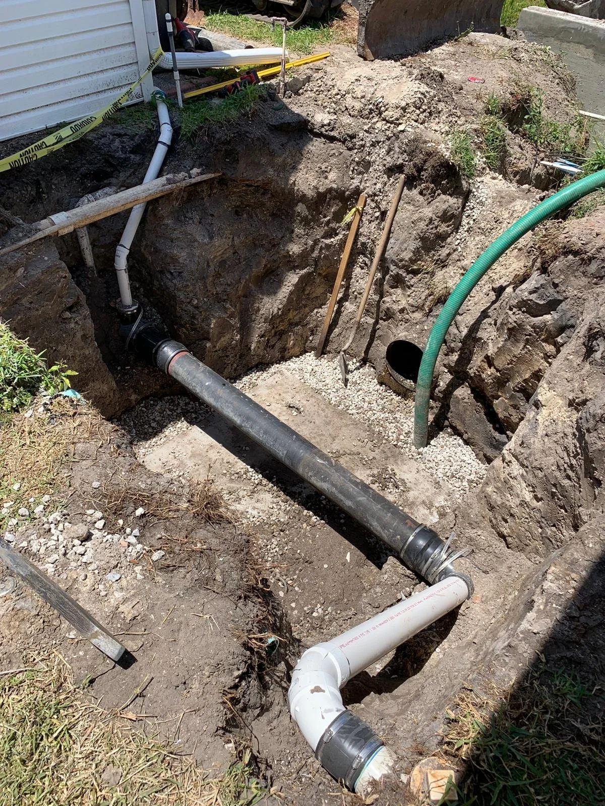 Excavation site with underground pipes, including black and white PVC pipes and a green flexible pipe, with construction tools and caution tape nearby. Utility conflict structure.