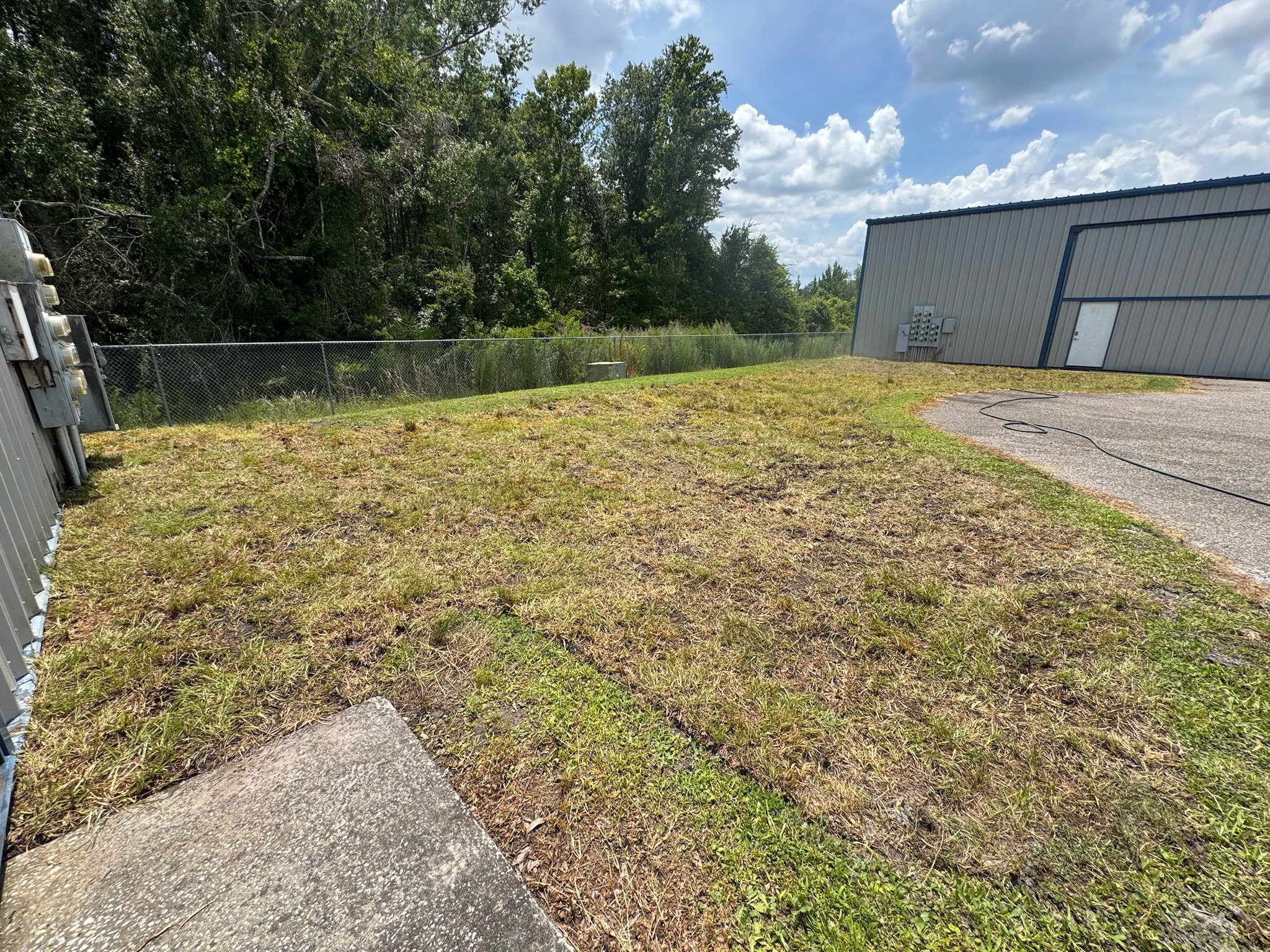 A grassy area next to a gravel pathway with a metal building on the right, gray with blue trim, and electrical panels on the side. A chain-link fence runs along the back, with trees and a partly cloudy sky in the background. Sod restoration.