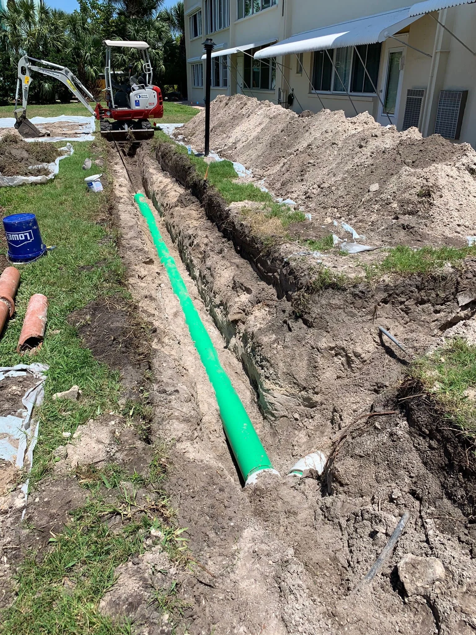 A construction site showing a trench being dug for underground plumbing, with a green pipe laid in the trench, a small excavator nearby, and dirt piles along the side of a building.