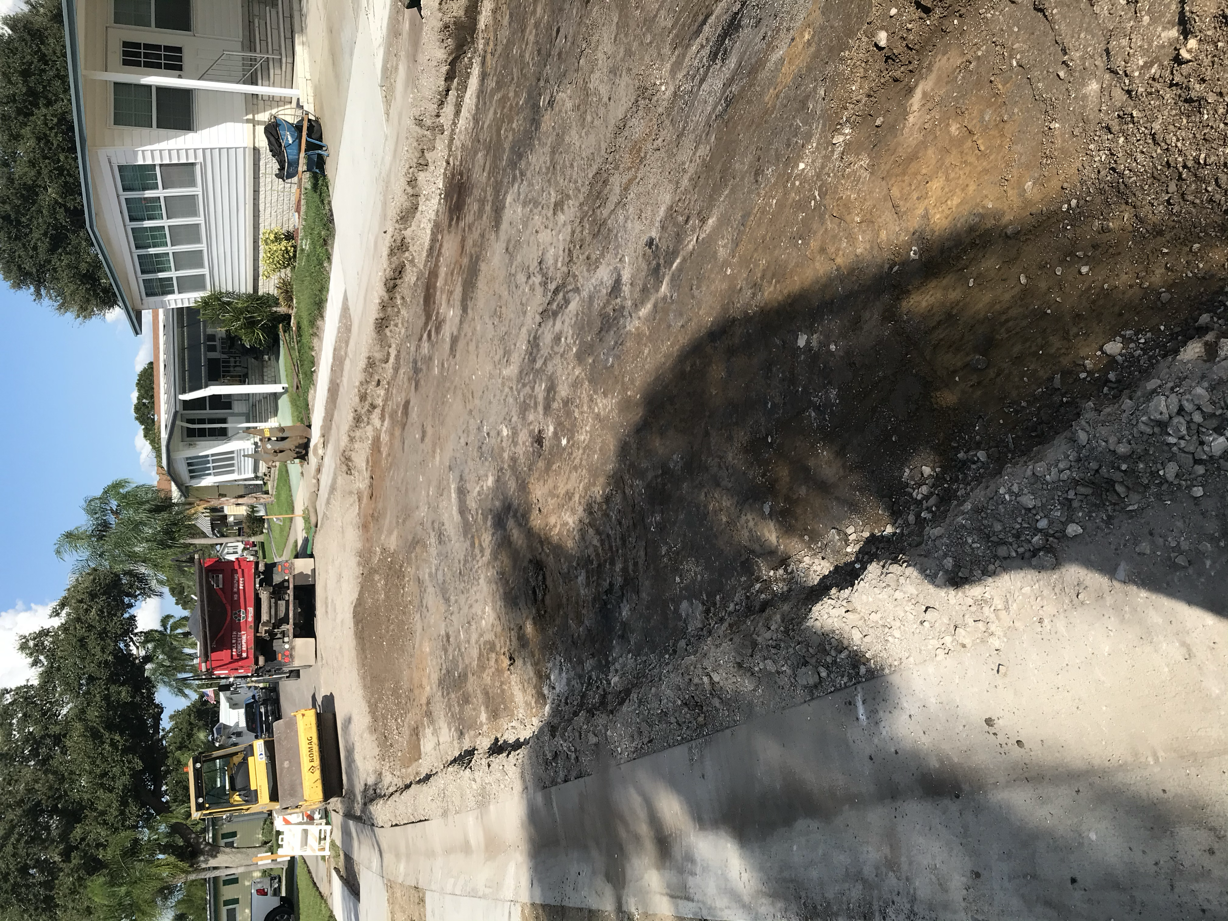 Construction site with an area of exposed dirt and construction equipment, including a red truck and a yellow excavator, in a residential neighborhood. Roadway reconstruction.