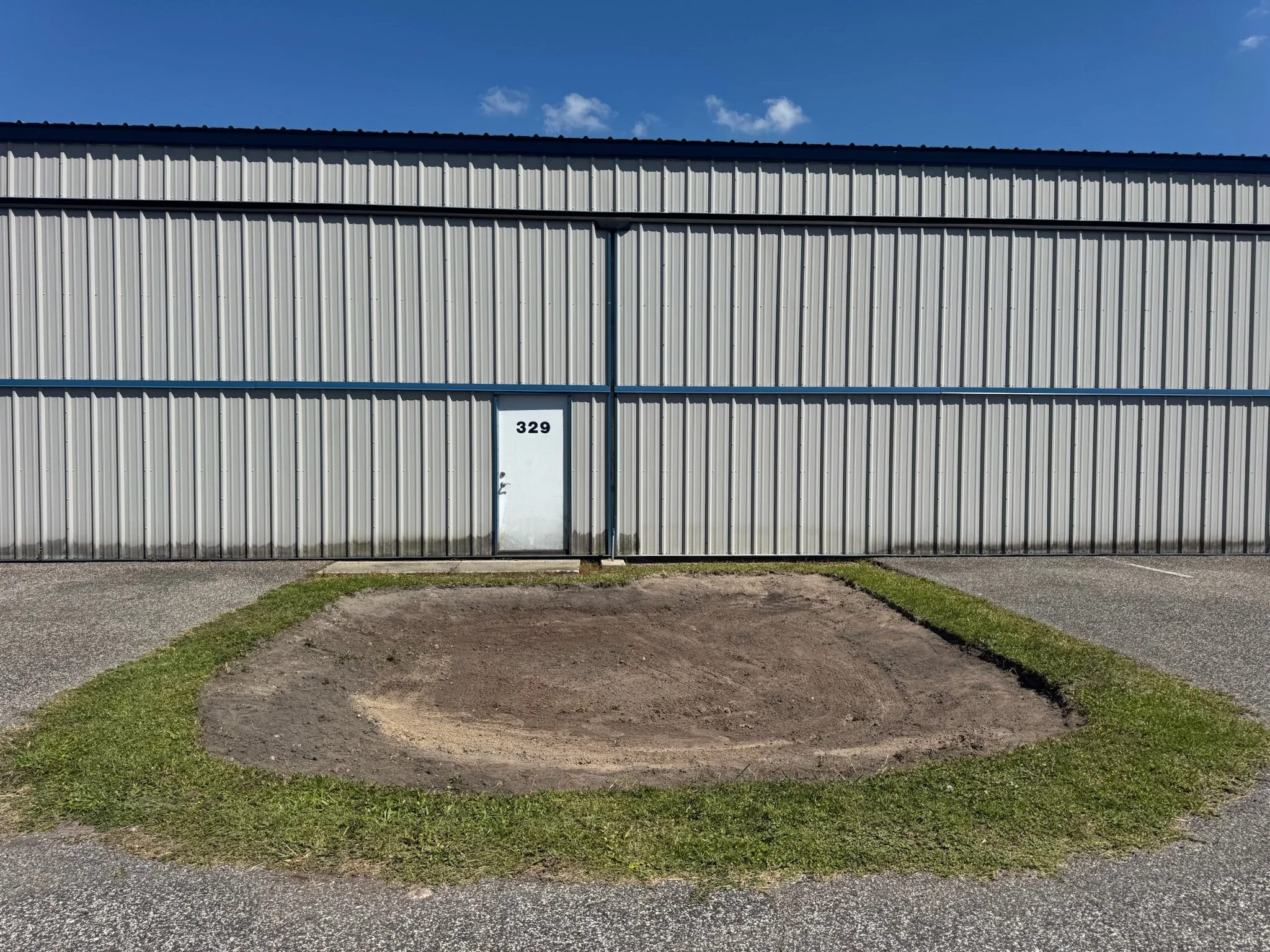 A metal building with a white door labeled 329, surrounded by a gravel area and patches of grass, under a blue sky with a few small clouds. Water retention area regrading.