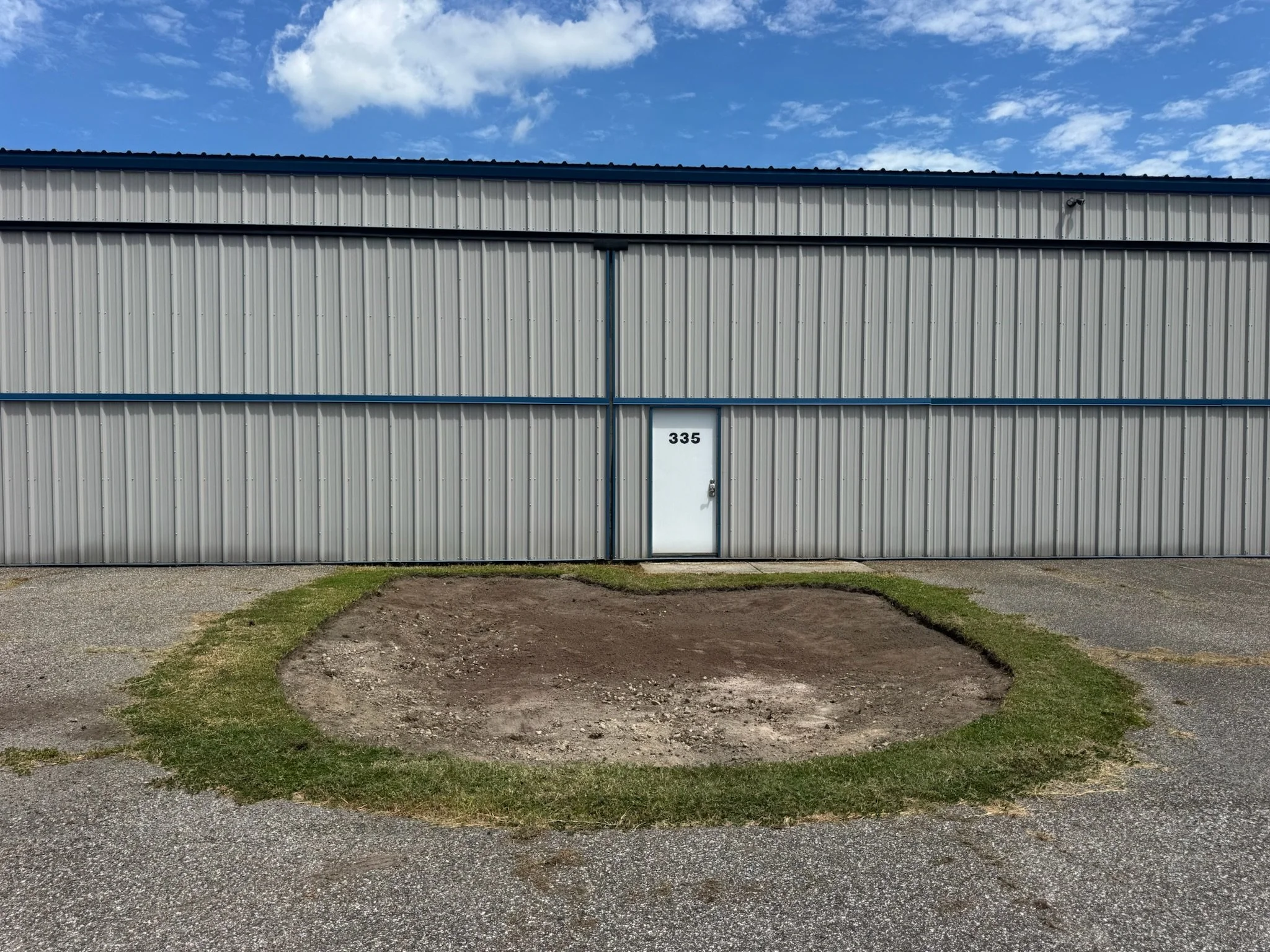 The exterior of a metal building. The building has horizontal siding with blue trim and is set against a partly cloudy sky. There is a dirt patch and some grass in front of the building. Elevation regrading for drainage improvements.