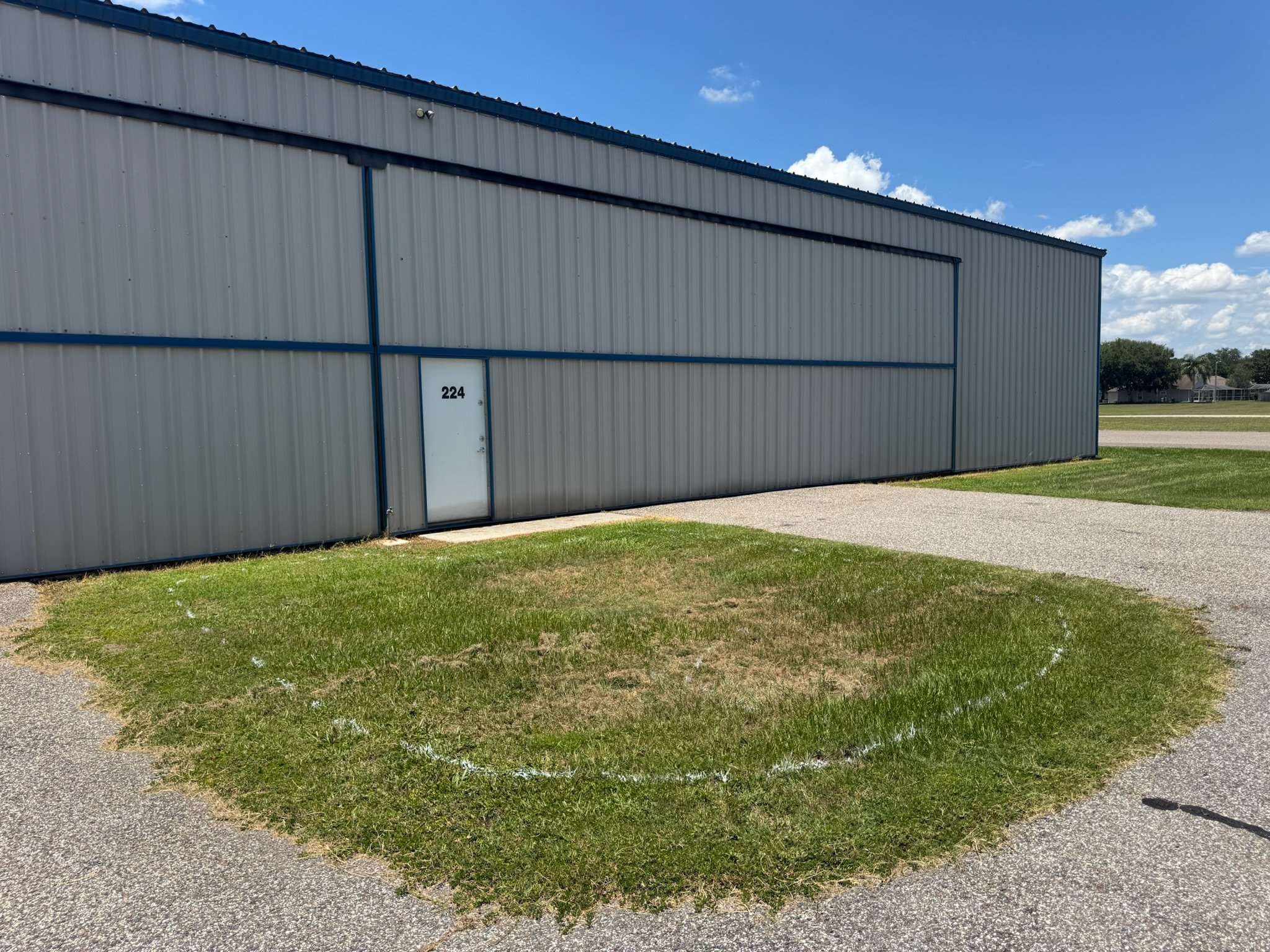 A metal building with a white door labeled 224, set against a blue sky with clouds. The building is situated in a grassy area with a concrete path in front and a residential neighborhood visible in the background. Grass removal prep.