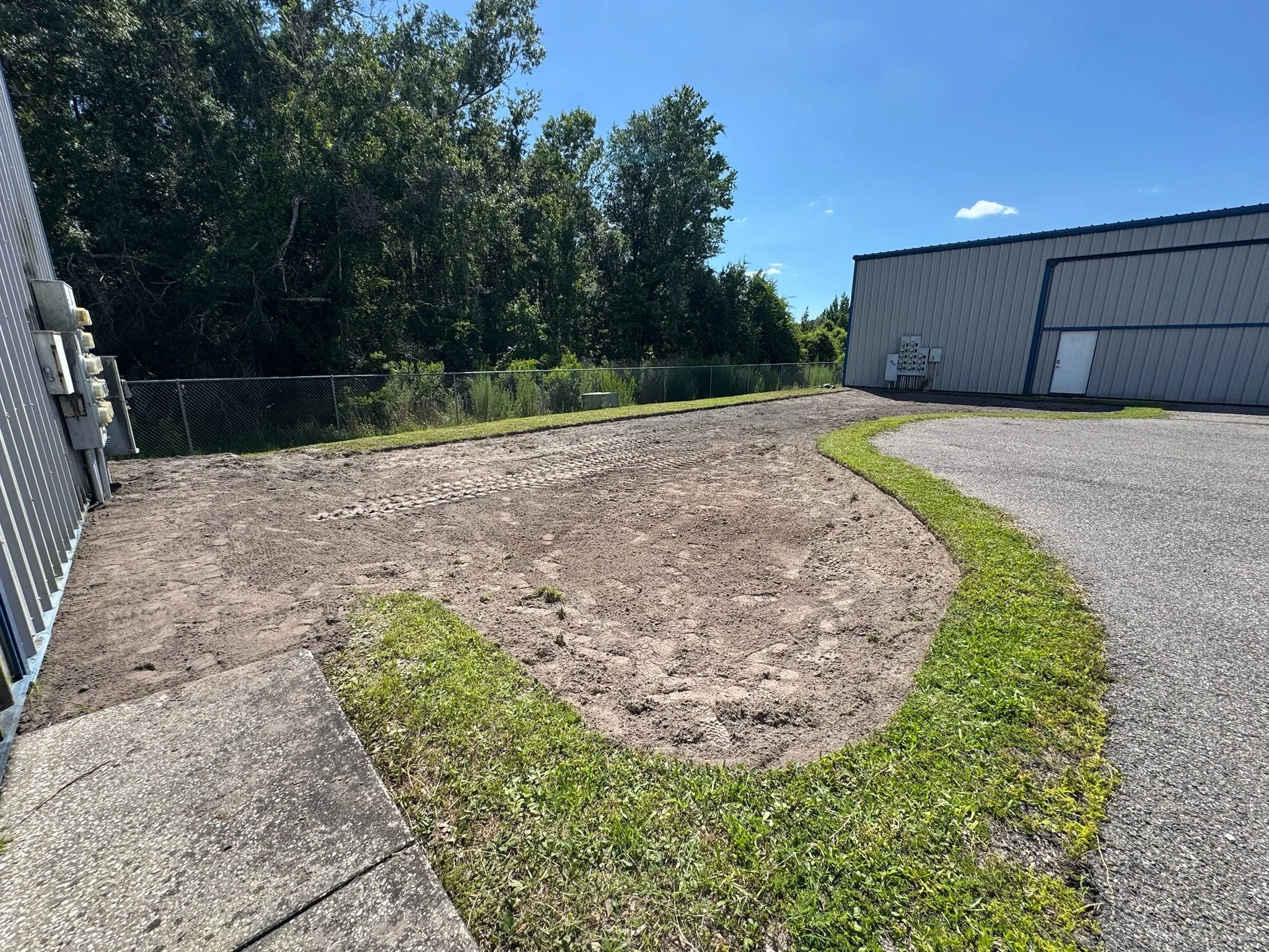 Outdoor scene with a gravel and concrete ground, a grassy patch, a building with metal siding on the right, a chain-link fence, trees and blue sky in the background. Elevation regrading for drainage.