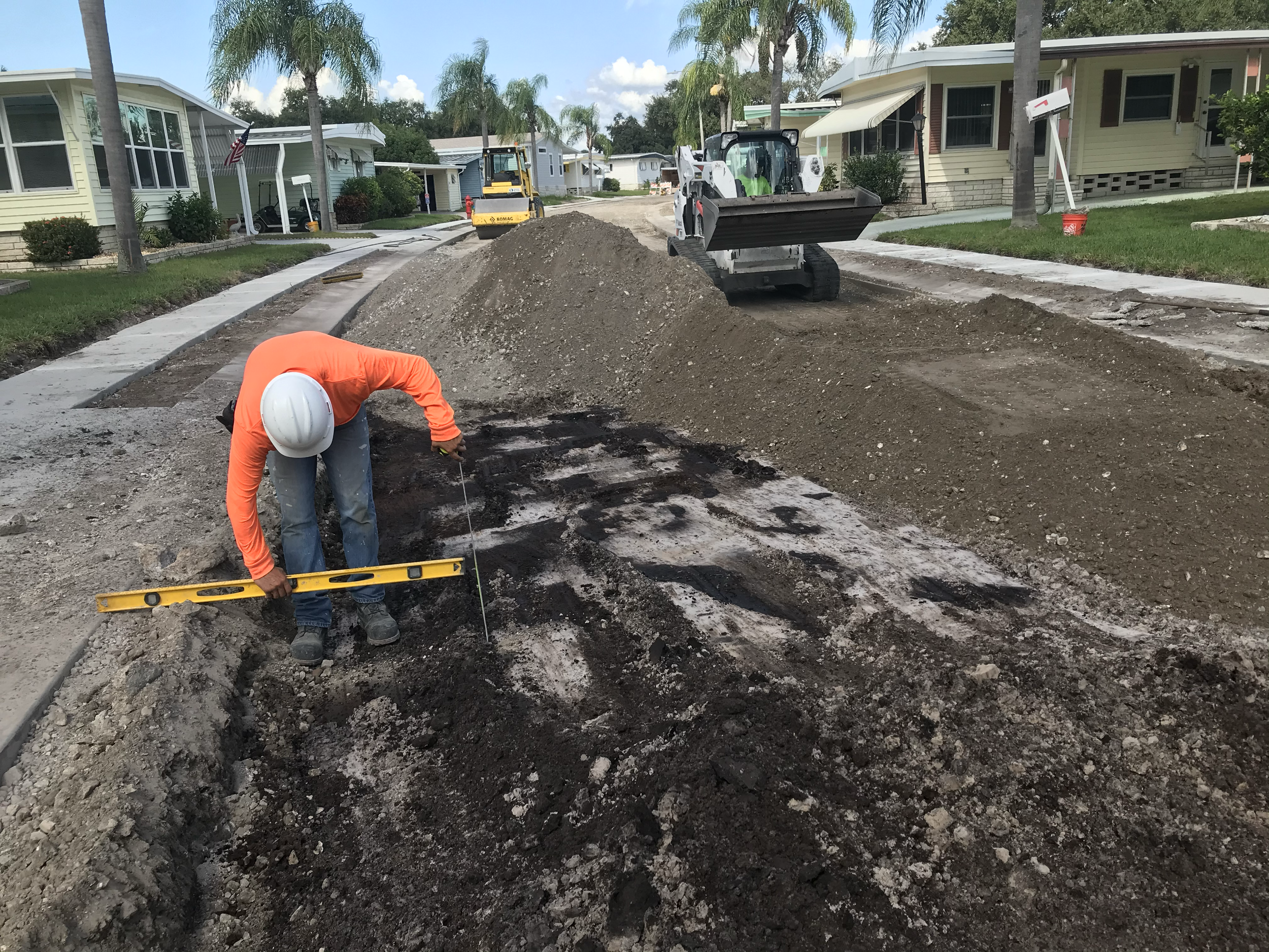 Construction workers in hard hats working on a street repair project with excavation equipment, surrounded by residential houses and palm trees in a neighborhood. Roadway reconstruction.