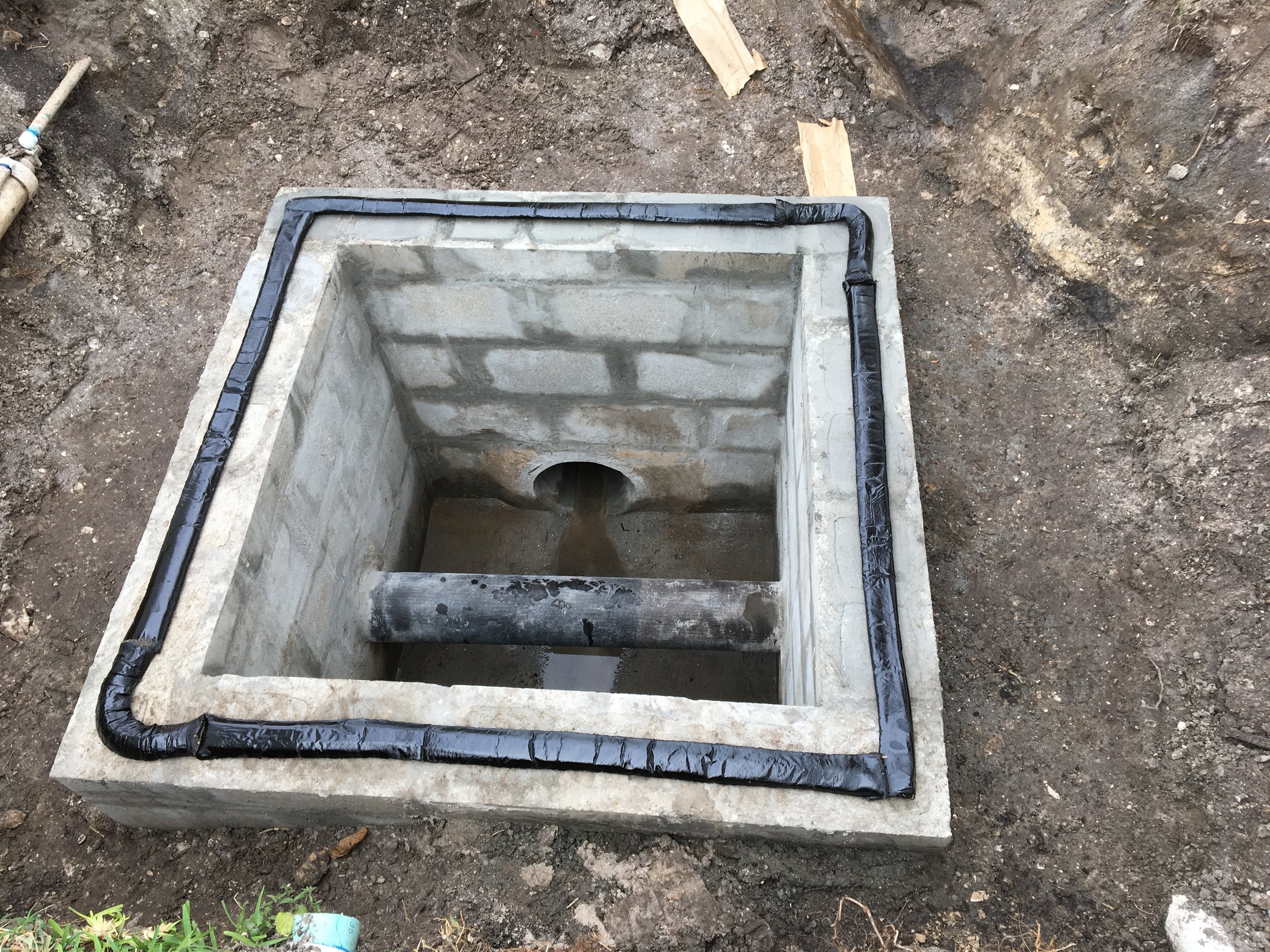 A concrete foundation or base with black waterproofing or drainage material installed around the edges, situated in an outdoor soil area. Utility conflict structure.