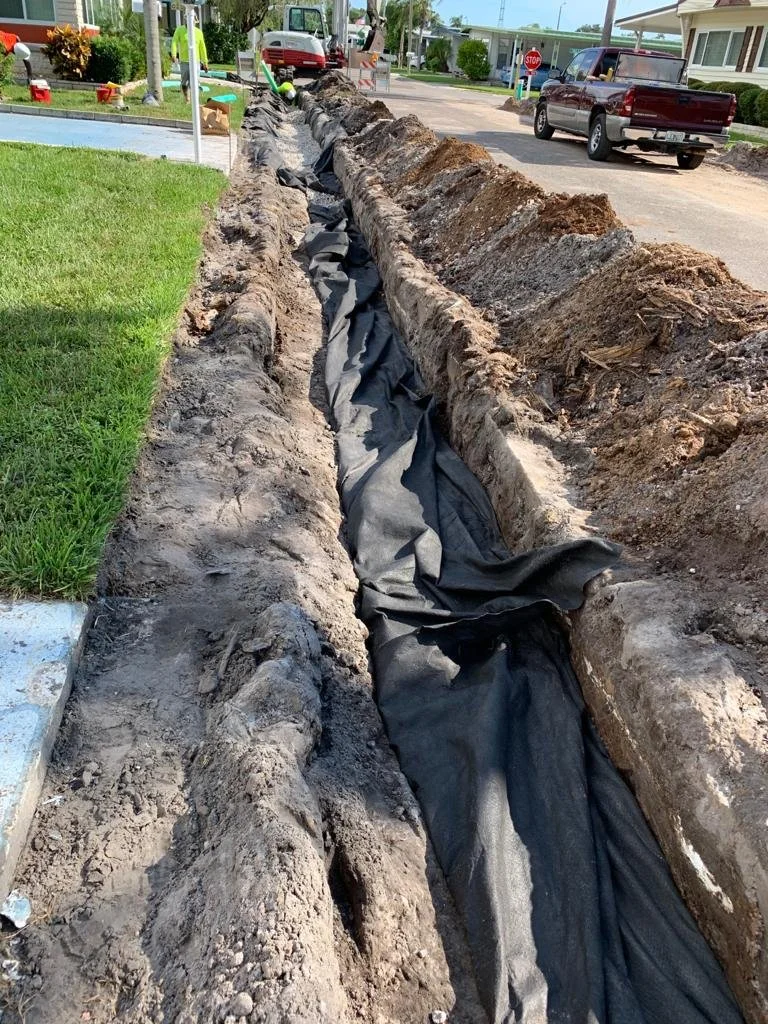 Construction workers laying underground piping in a dug trench along a street, with a truck and cars in the background.