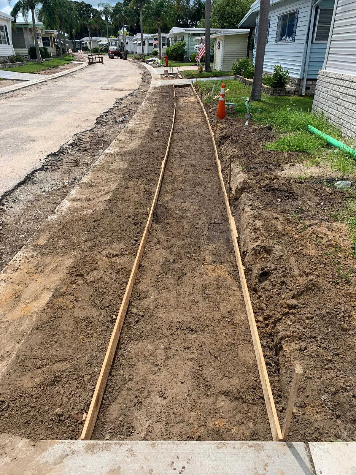 Construction of a new sidewalk or pathway with wooden forms and dirt, in a residential neighborhood with houses, trees, and construction tools and cones.