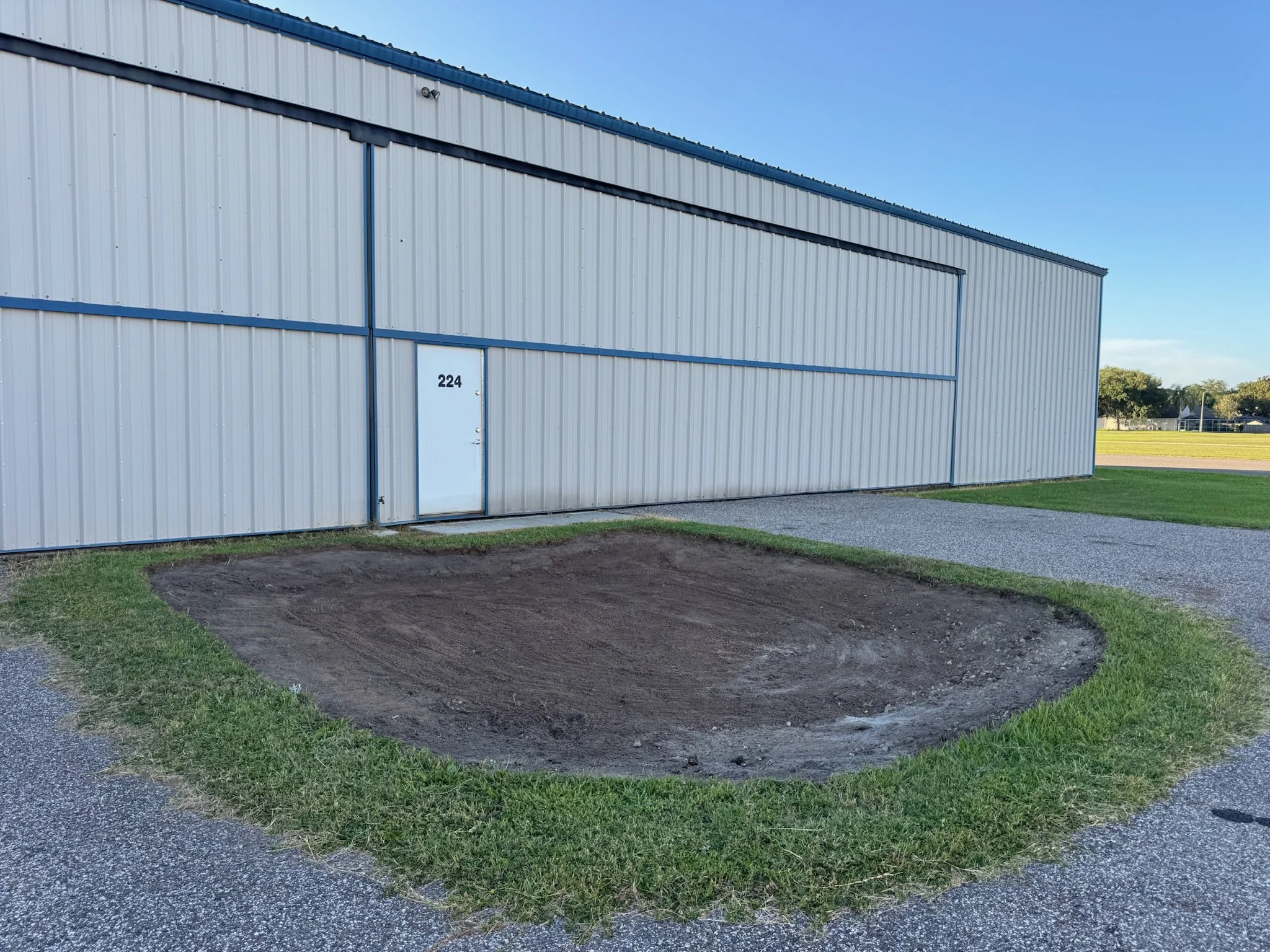 A large metal building with a blue trim and the number 224 on the door, situated on a grassy area with a gravel pathway and some trees in the background under a clear blue sky. Water retention area regrading.